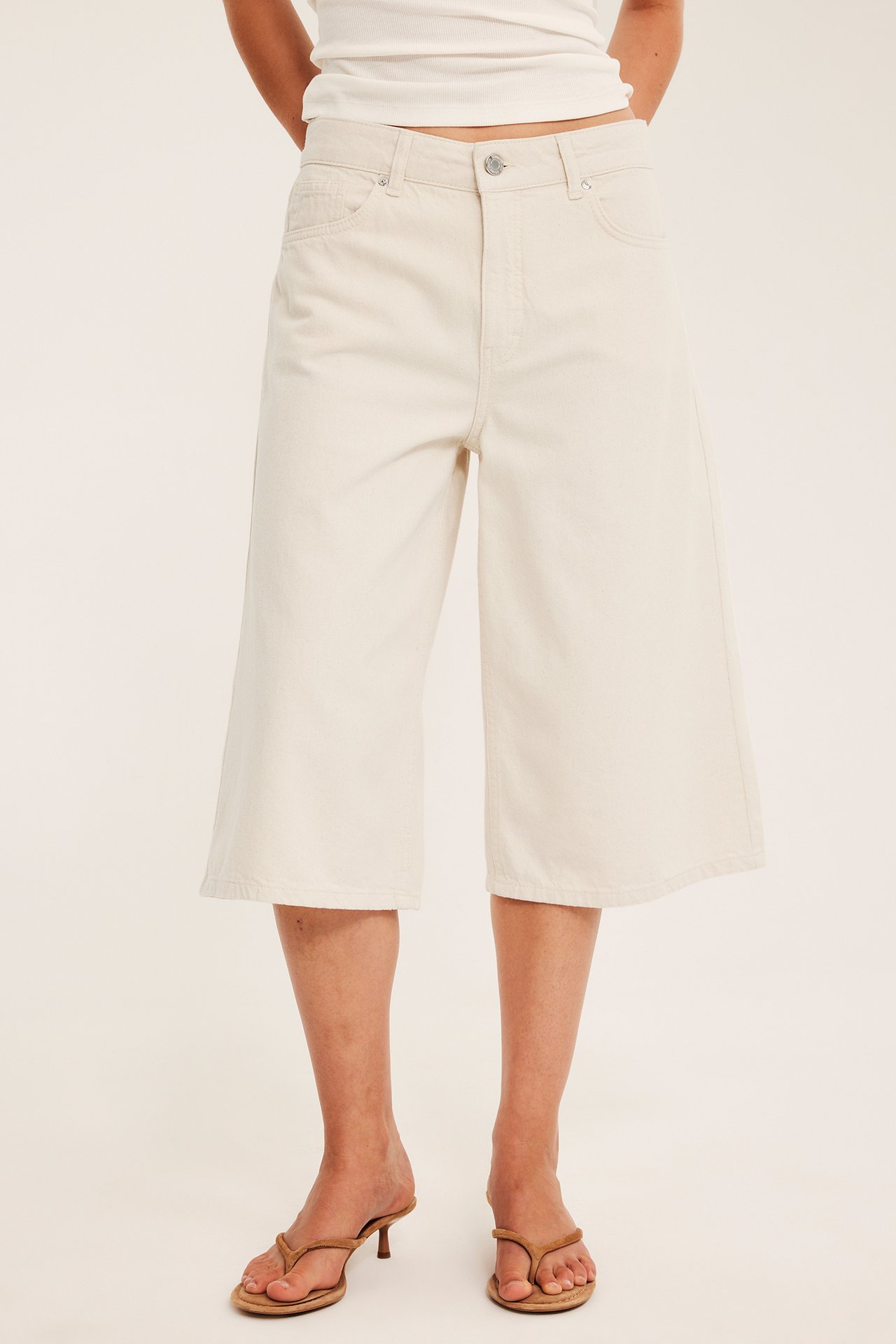 Edestä: nainen valkoisissa löysissä puuvillatwill-bermudashortseissa, valkoinen toppi ja ruskeat sandaalit.