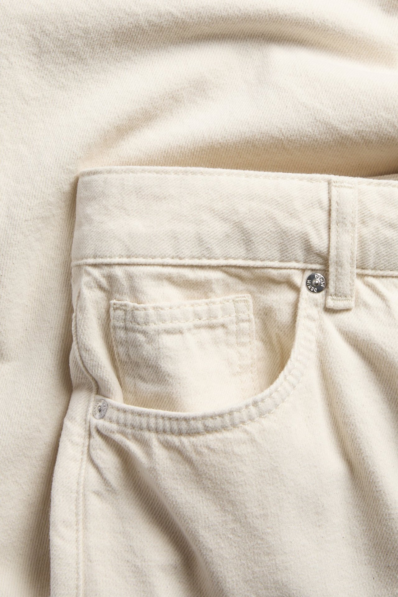 Närbild, framifrån, av vita bomullstwill-bermudashorts med fem fickor och metallknapp.