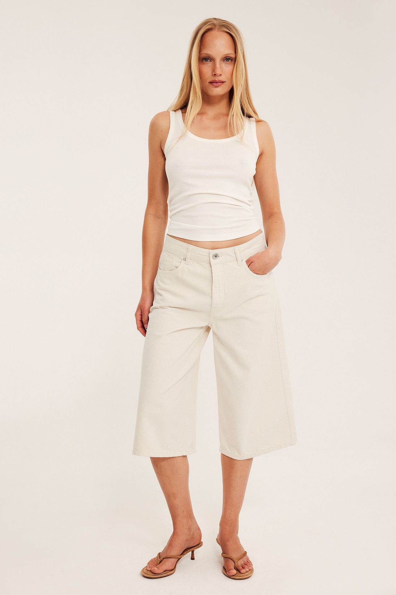 Nainen edestäpäin yllään valkoiset löysät puuvillatvillibermudashortsit, valkoinen ribbitoppi ja ruskeat korkokengät.