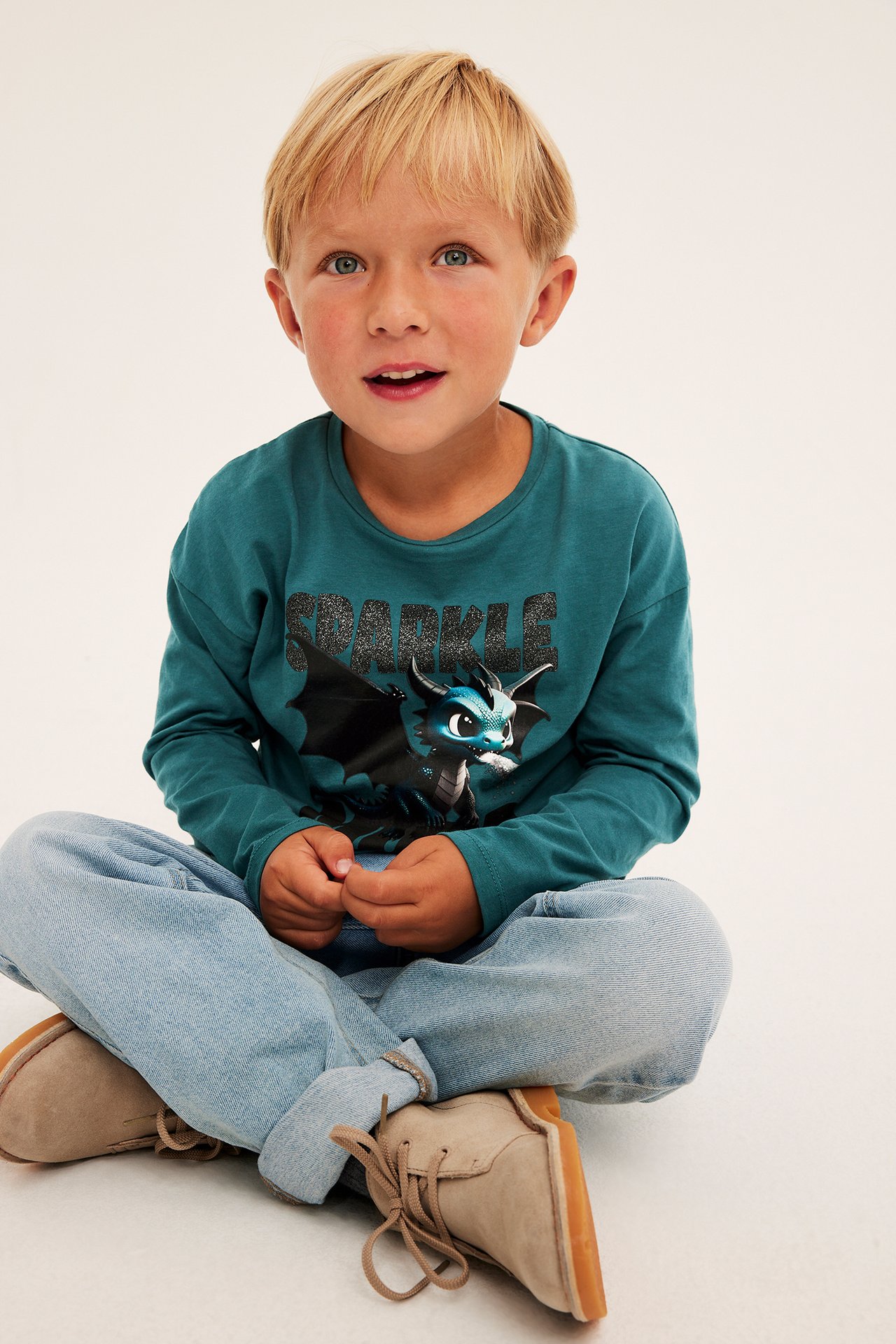 Frontvy av ett barn i mörkblå långärmad t-shirt med Sparkle-drakprint, ljusblå jeans och beiga kängor.