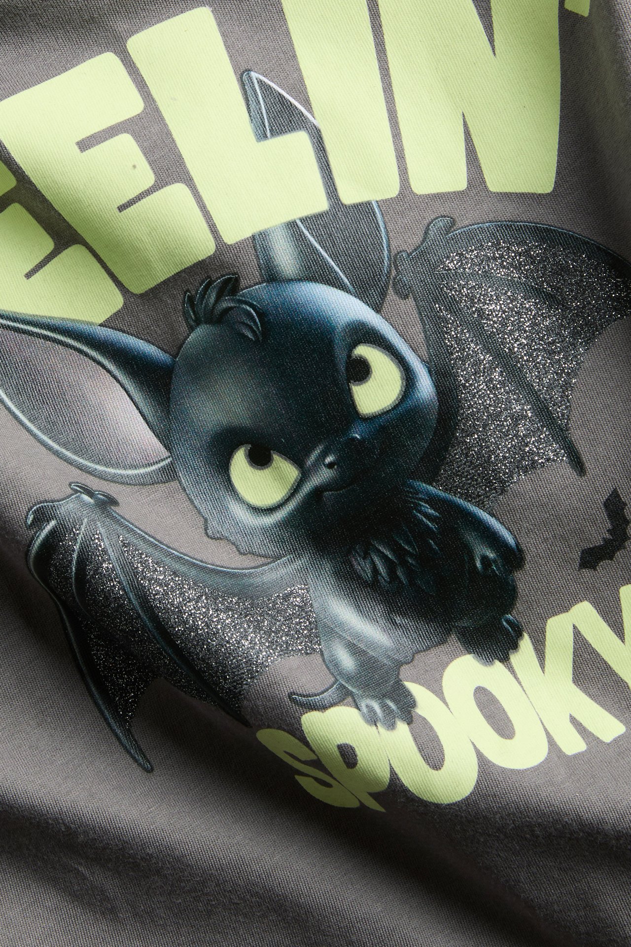 Närbild på grå t-shirt med svart fladdermusliknande tryck, glödande ögon, glittervingar och texten 'SPOOKY'.
