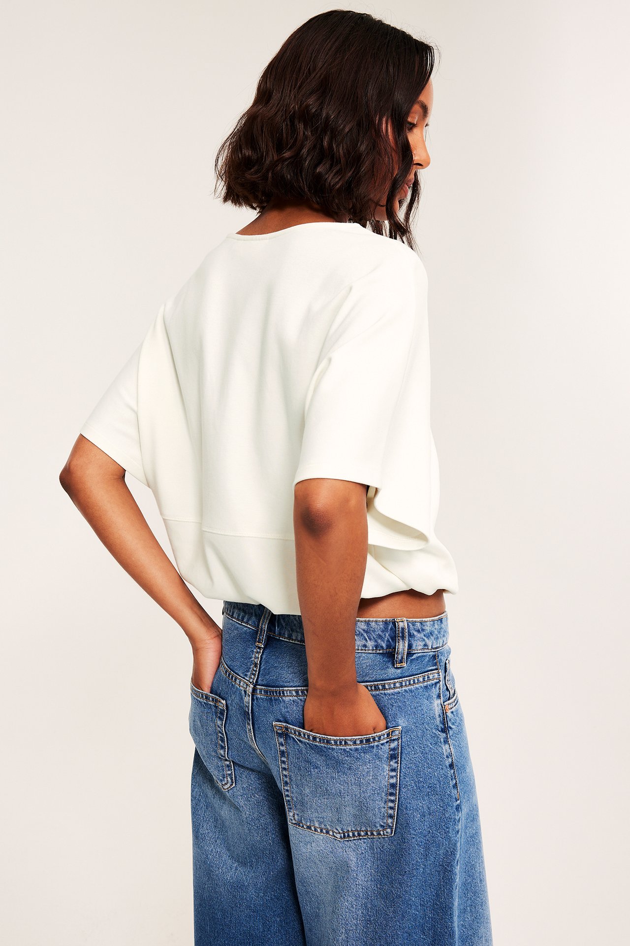 Kobieta tyłem w kremowym, luźnym topie z krótkim rękawem i niebieskich jeansach wide-leg.