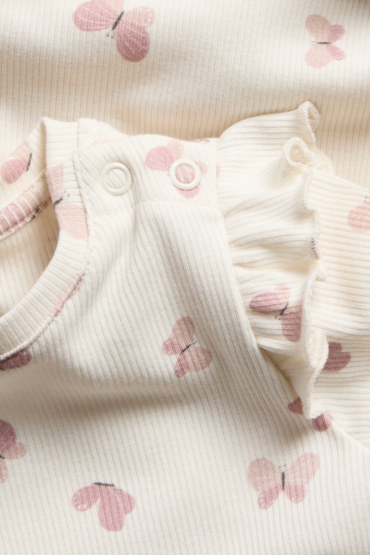 Närbild av en ljusrosa ribbad babybody med volangaxlar och rosa fjärilsmönster, med tryckknappar.