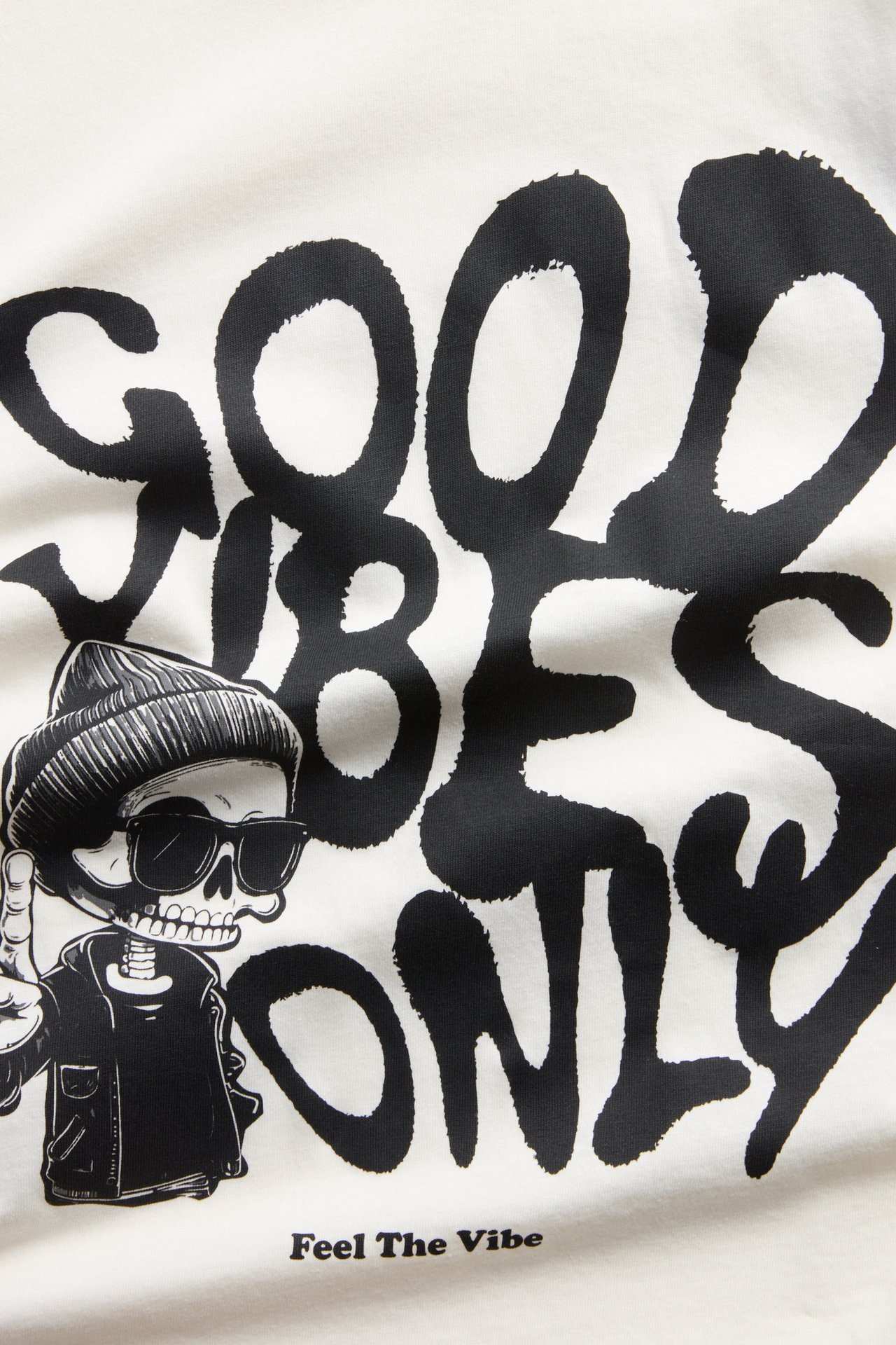 Närbild på en vit topp med svart 'GOOD VIBES ONLY' grafiskt tryck och skelettfigur.
