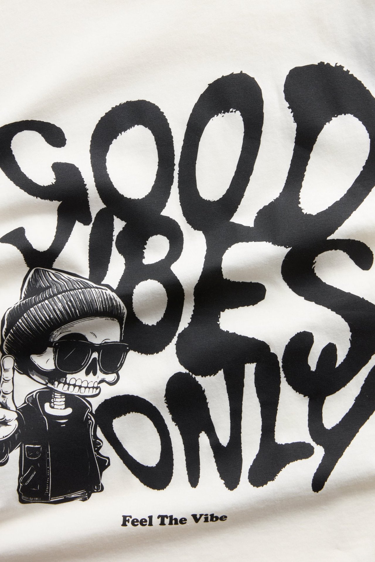 Närbild på en vit topp med svart 'GOOD VIBES ONLY' grafiskt tryck och skelettfigur.