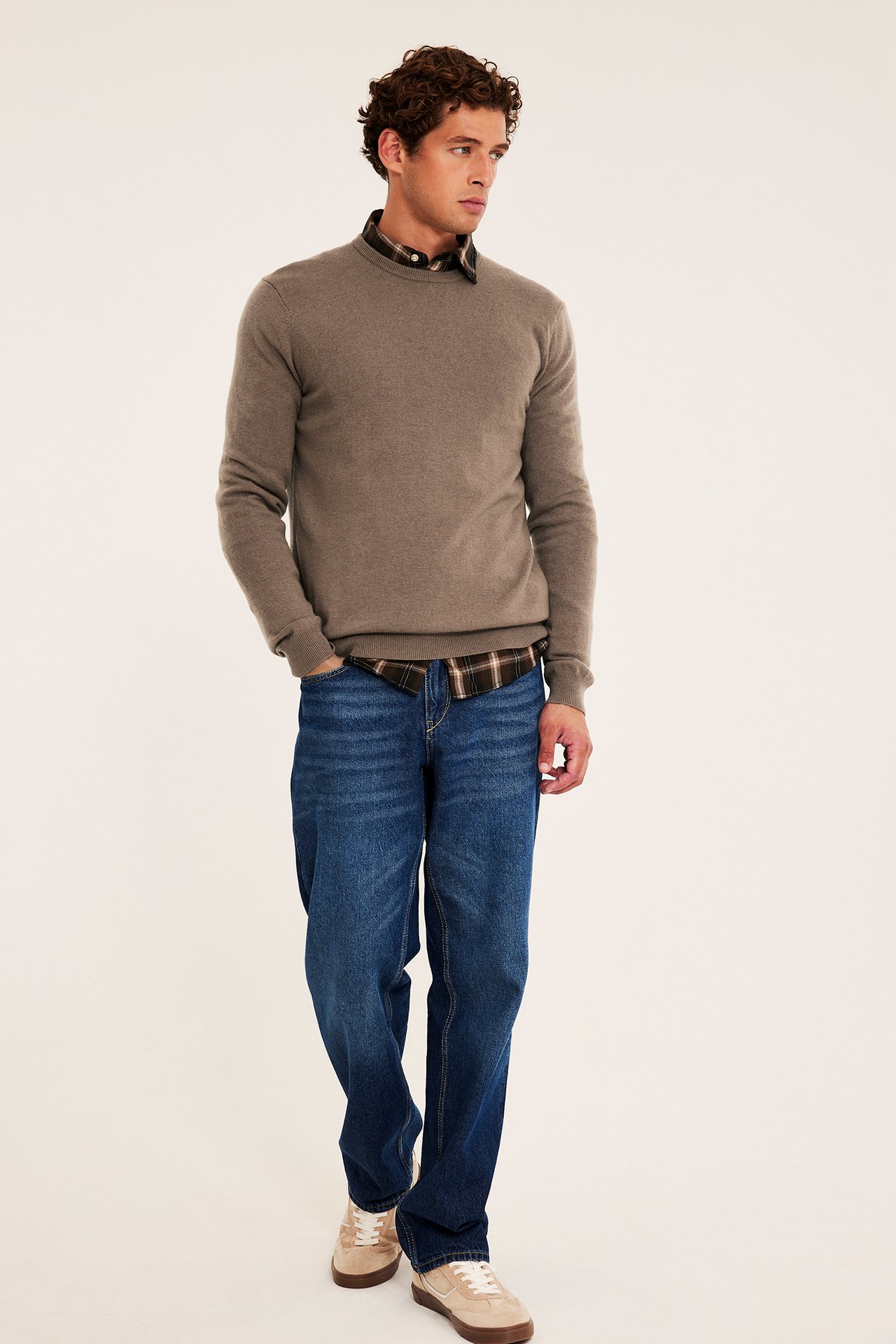 Frontbilde av mannlig modell i brun genser, rutete skjorte, blå jeans og beige joggesko.