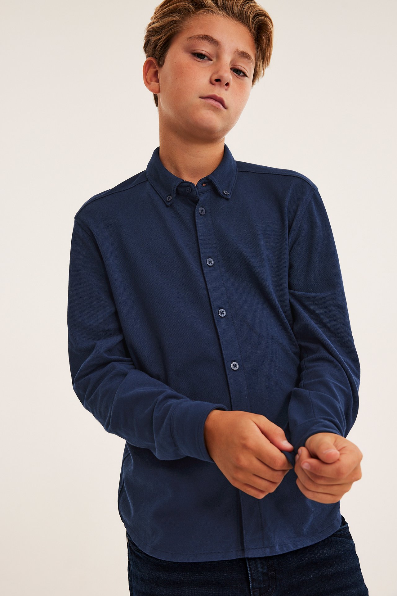 Framifrån, barn i marinblå långärmad skjorta med knappar, justerar manschett, och mörkblå jeans.