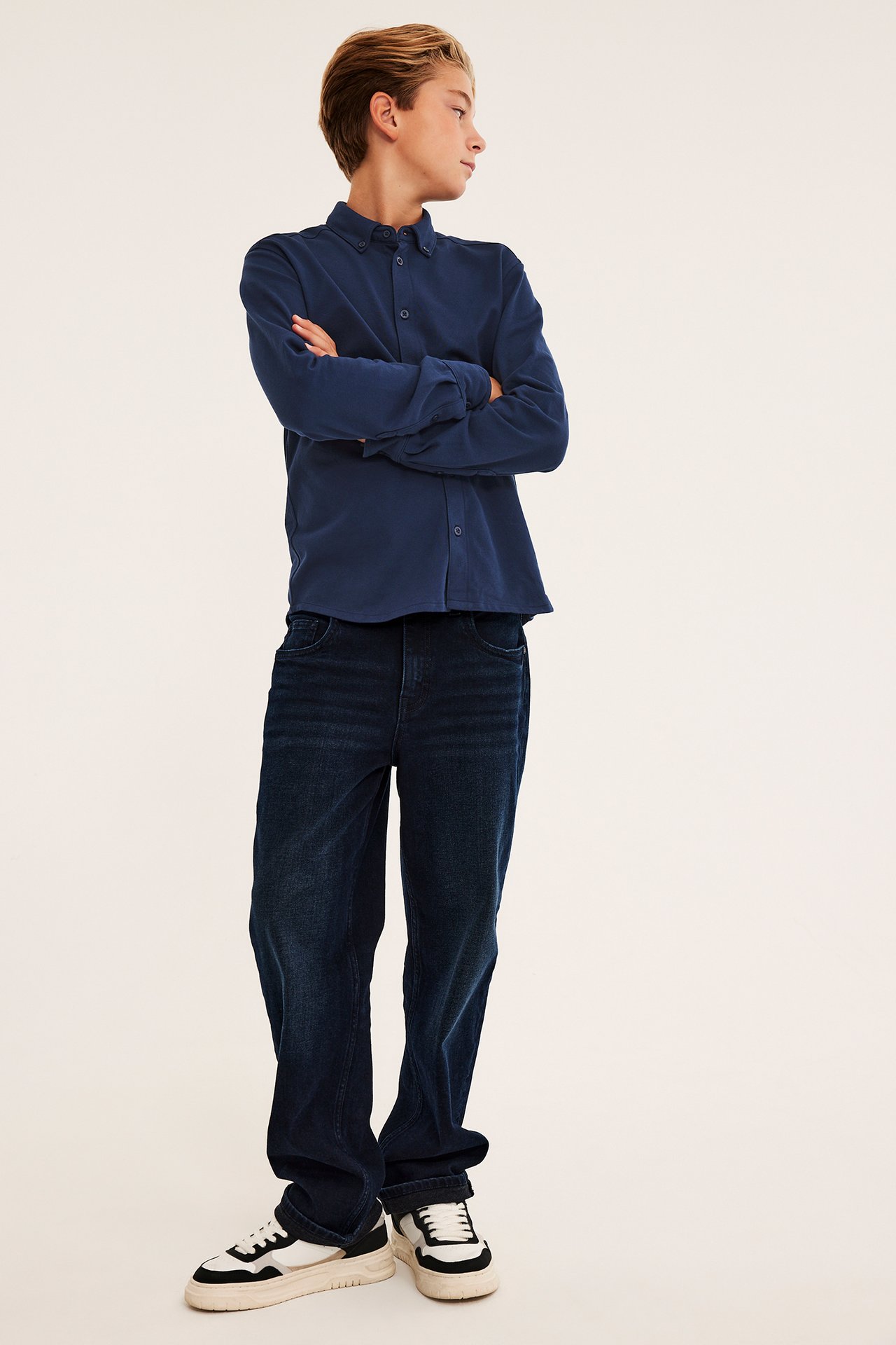 Frontvy av en pojke i mörkblå långärmad skjorta, mörkblå jeans och sneakers.