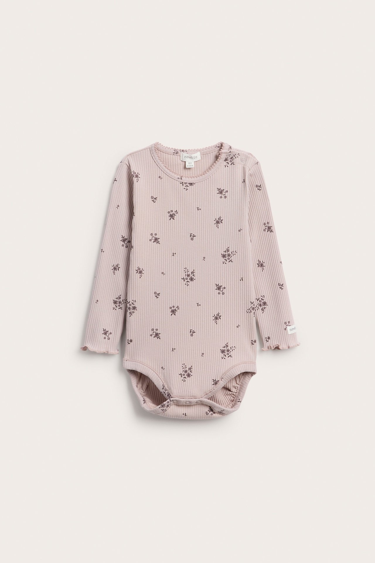 Frontvy av en ljusrosa ribbad långärmad babybody med mörkt blommigt mönster och volangdetaljer.