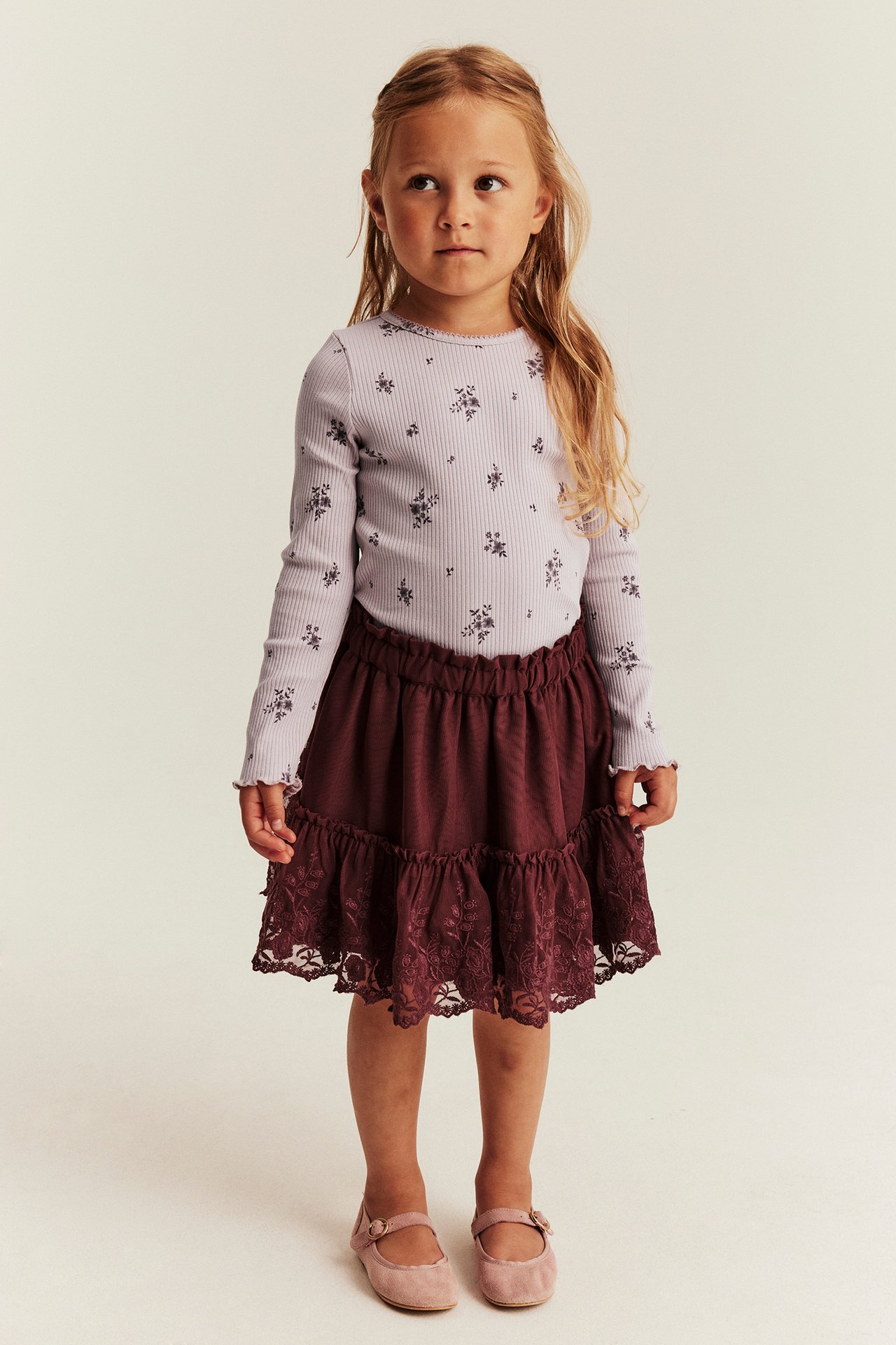 Widok z przodu: dziecko w jasnofioletowym kwiecistym topie, bordowej koronkowej spódnicy i różowych butach Mary Jane.