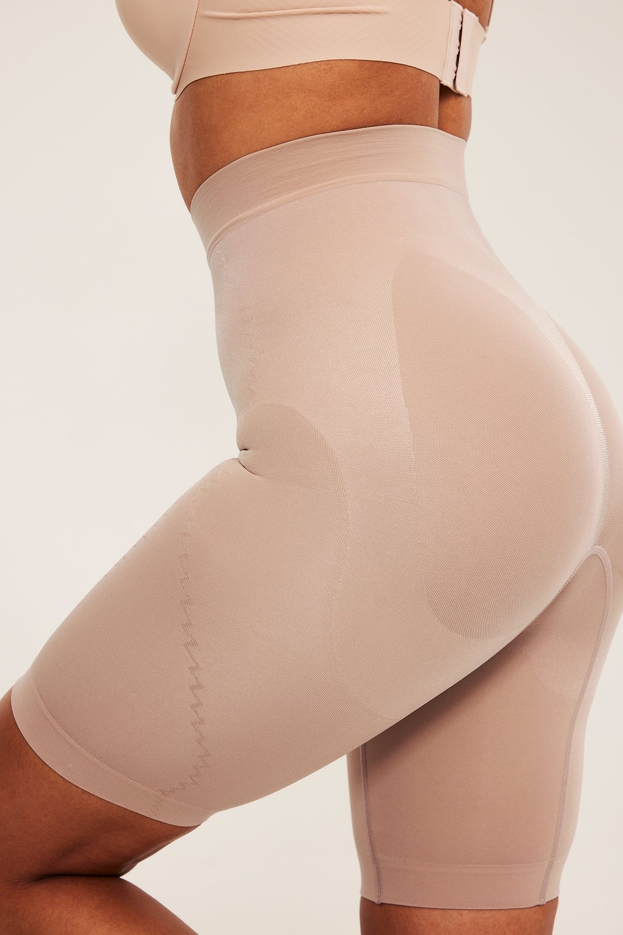 Sidebilde av en kvinne i beige sømløs shapingshorts med høy støtte og en beige BH.
