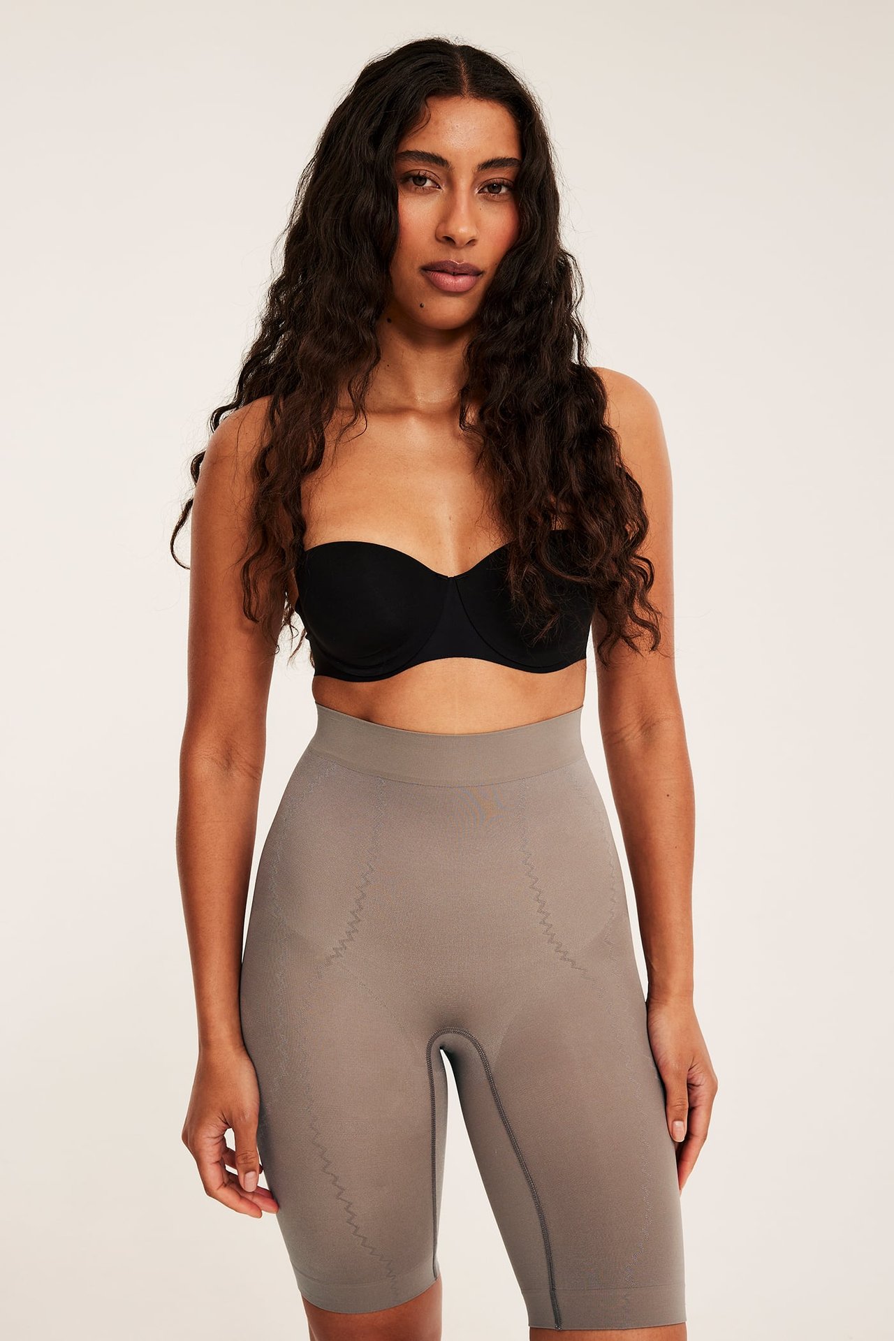 Framifrån, kvinna i seamless bruna högmidjade shapingshorts med vävda mönster och en svart axelbandslös BH.