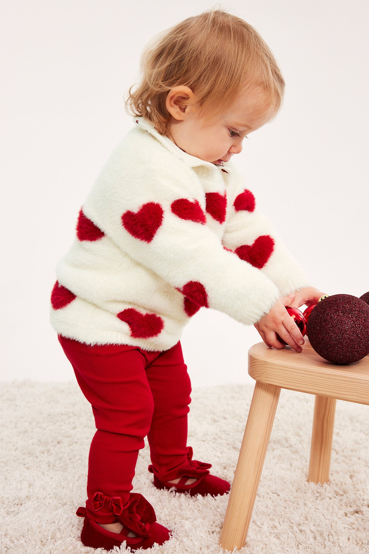 Sidovy av baby i vit, lurvig hjärttröja, röda ribbade leggings och röda sammetsskor.