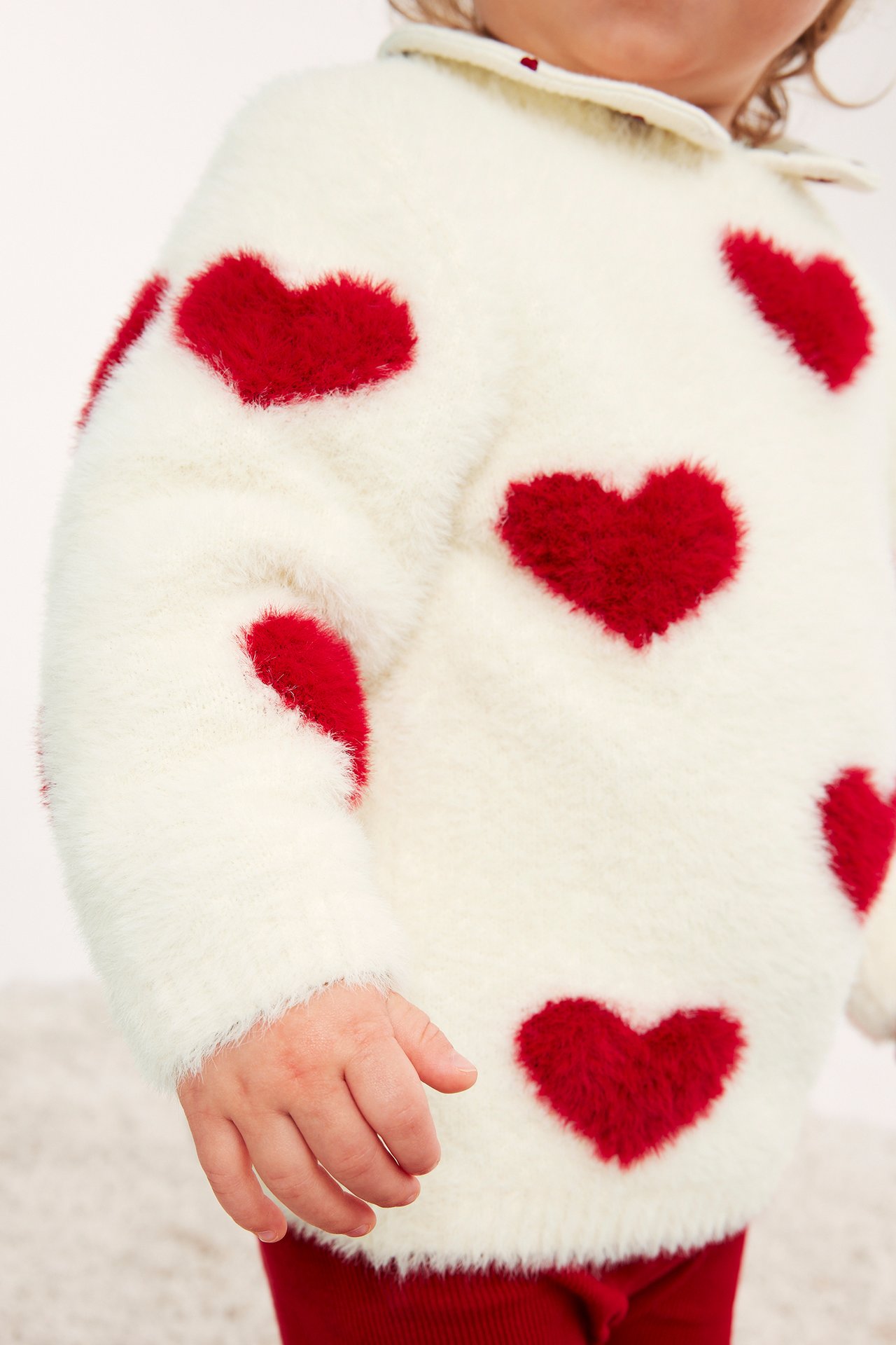 Närbild på baby i krämfärgad fluffig tröja med röda hjärtan och röda ribbade byxor.