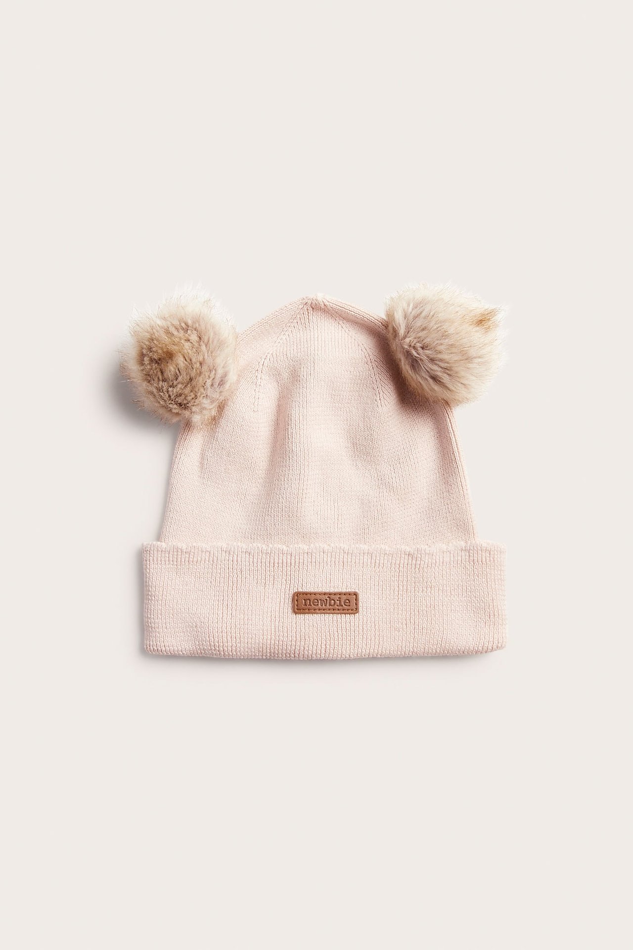 Front, lys rosa strikket babylue med to fuskepelsdusker og et brunt merke.