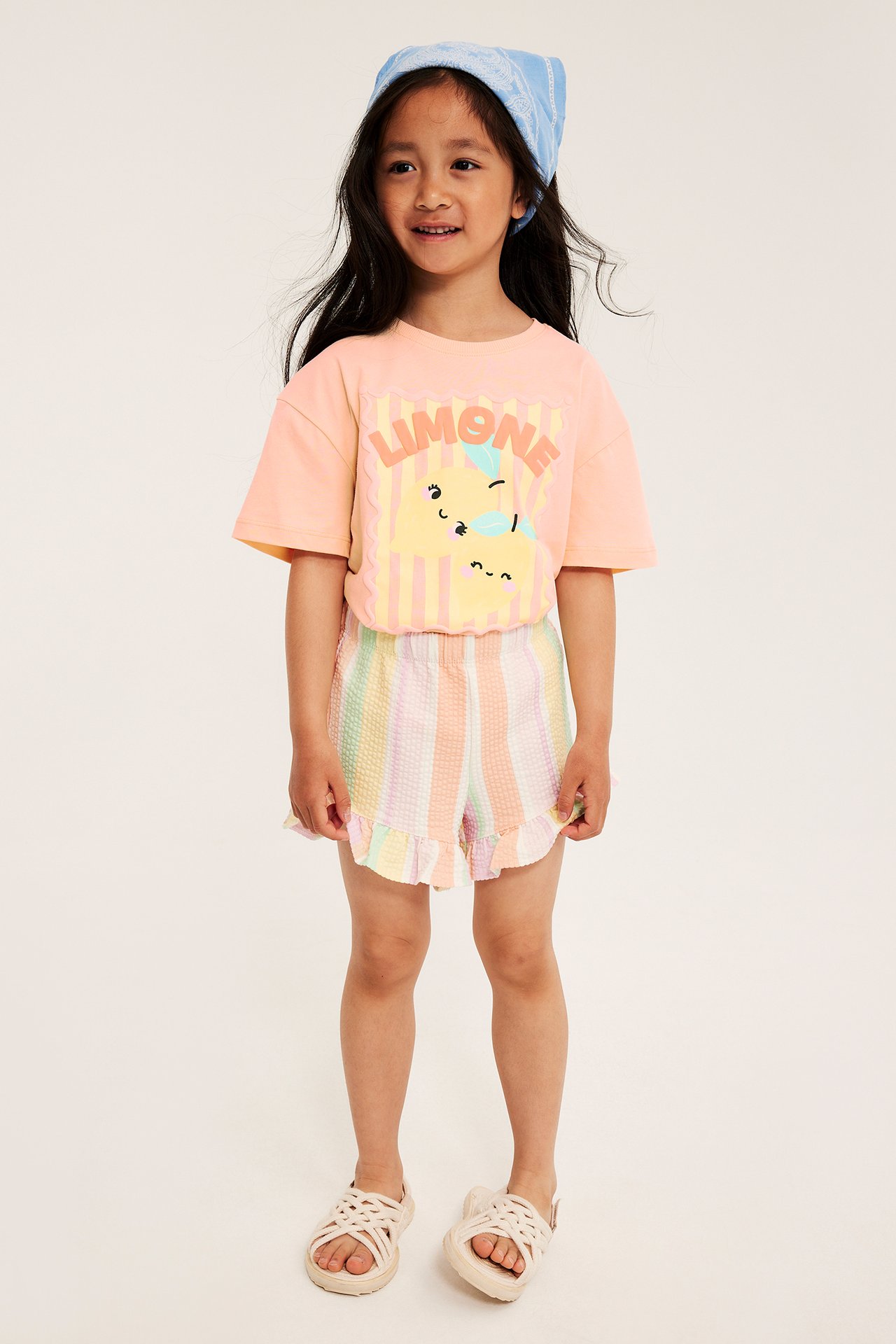Kid bär en korallfärgad bomulls-T-shirt med citronmönster, pastellrandiga shorts, blå bandana och beiga sandaler.