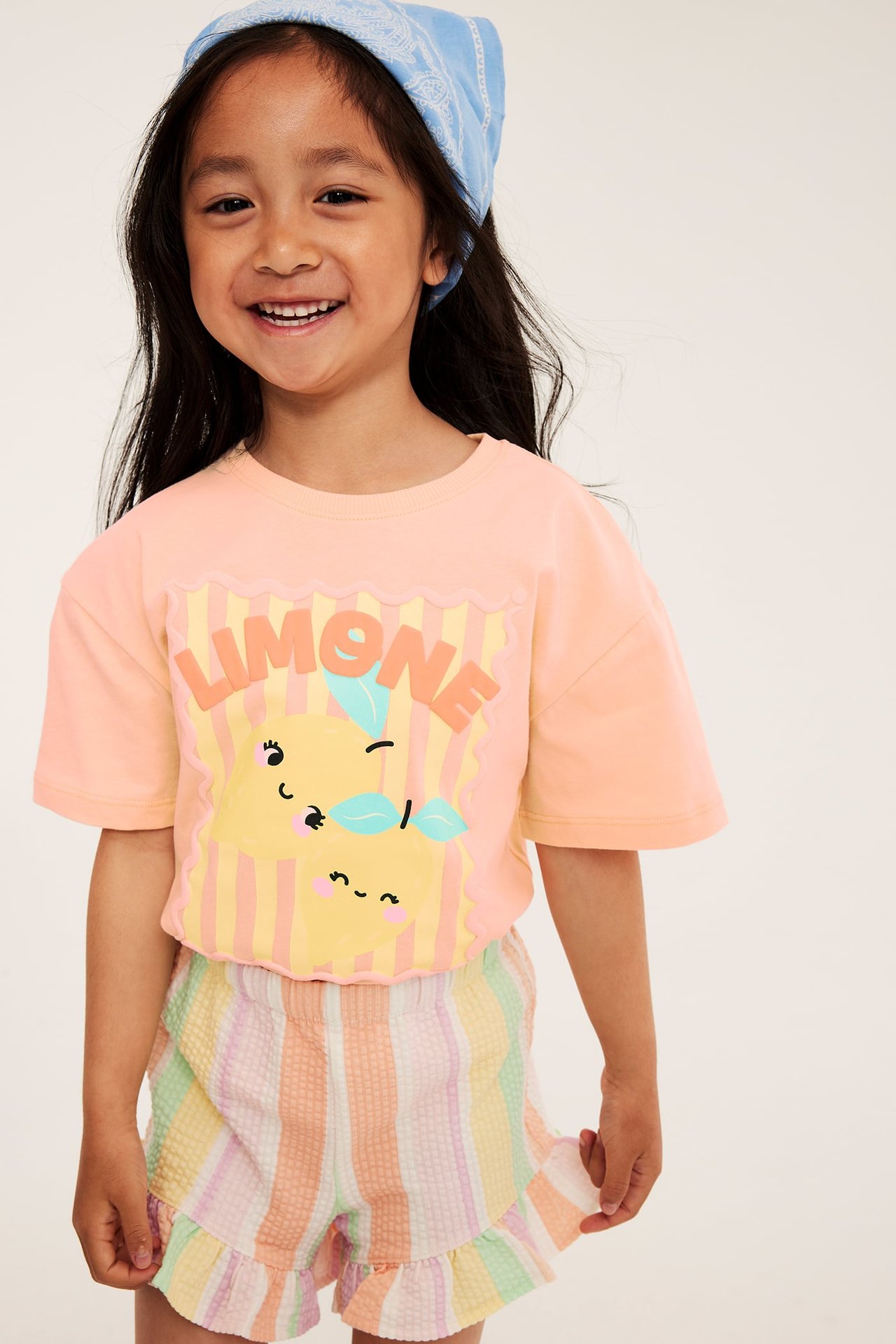 Framifrån: Barn bär korallfärgad t-shirt med citronmönster, pastellrandiga shorts och en blå bandana.