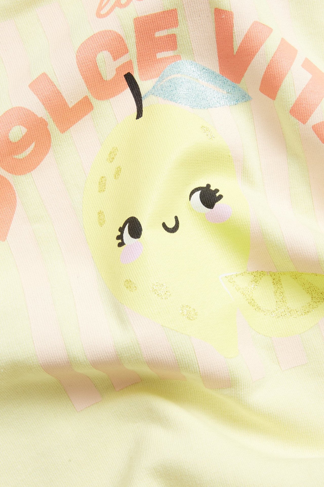 Nærbilde: lys gul genser med søtt sitronmotiv, tekst, glitterdetaljer og rosa striper.