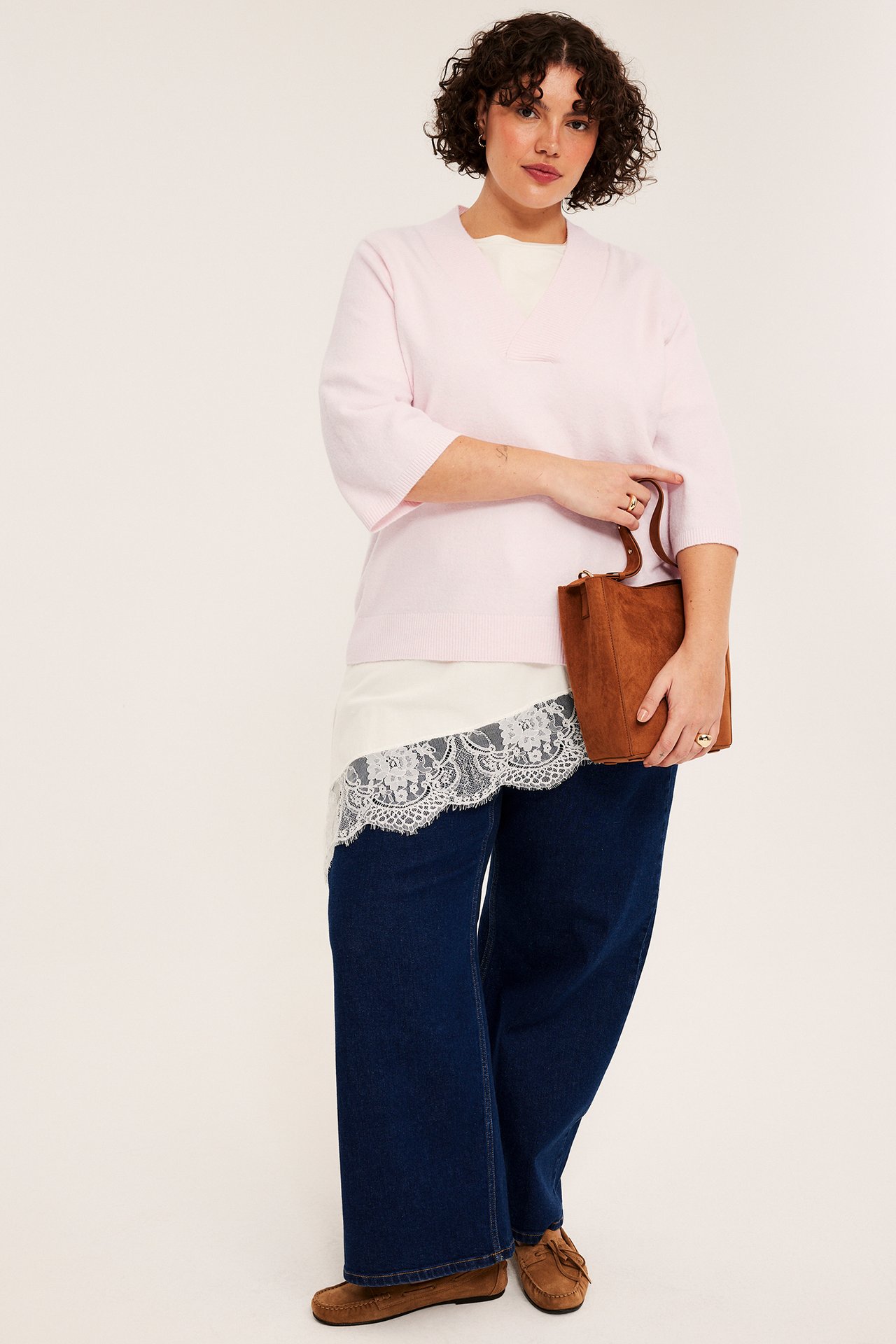 Kvinna i ljusrosa v-ringad tröja, vit spetstopp, mörkblå jeans, bruna mockasiner, håller brun väska.
