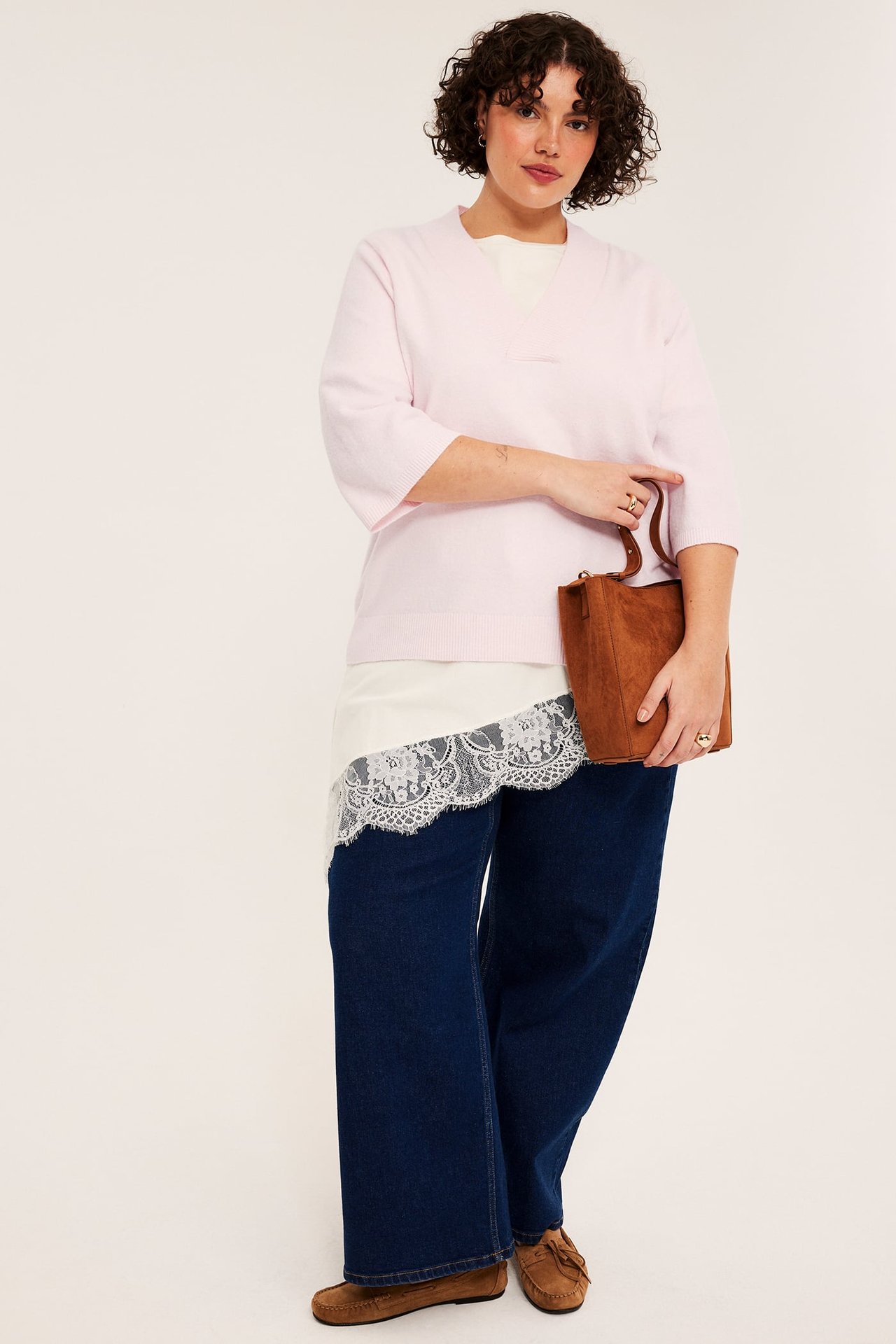 Kvinna i ljusrosa v-ringad tröja, vit spetstopp, mörkblå jeans, bruna mockasiner, håller brun väska.