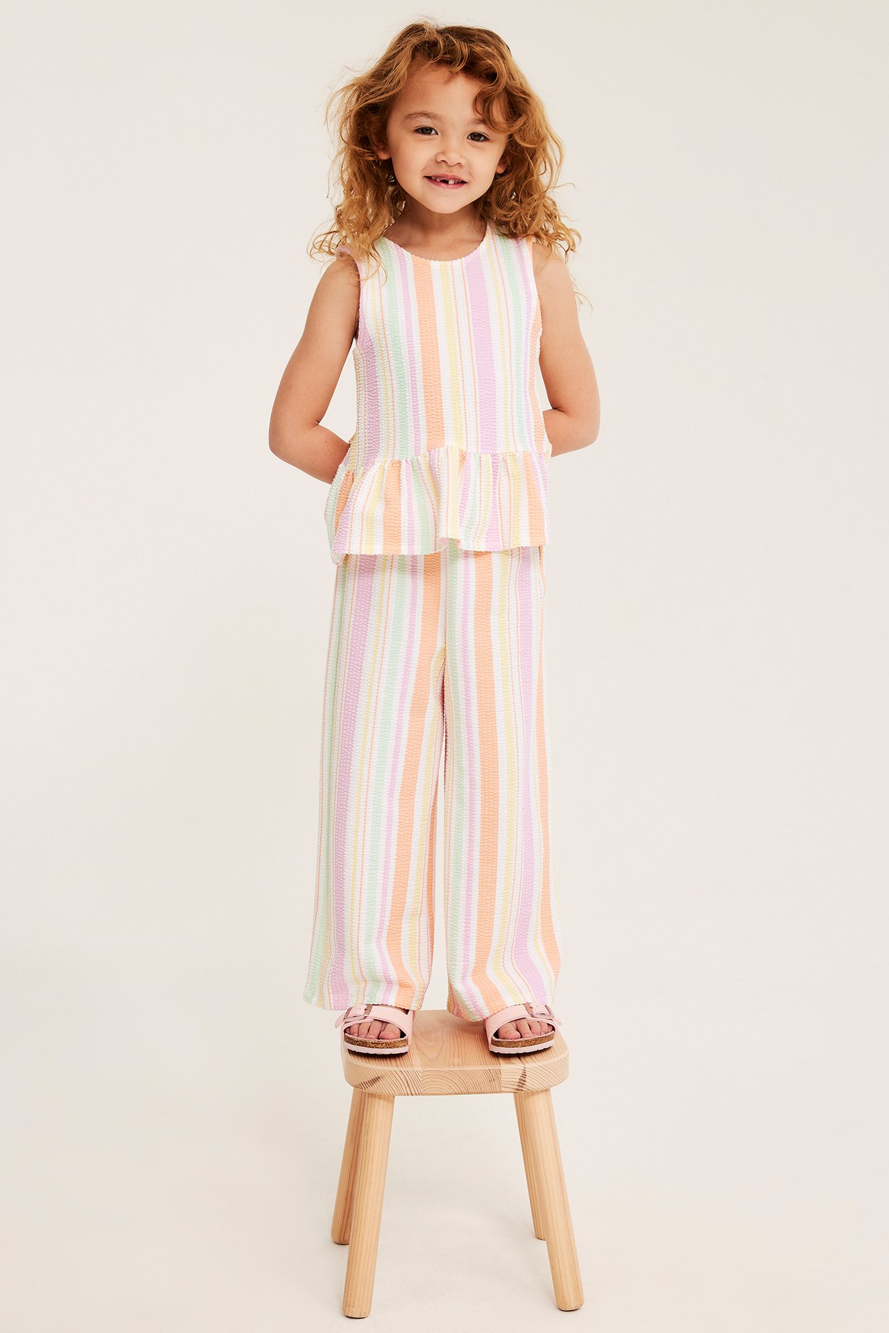 Widok z przodu: Dziecko w pastelowych, pasiastych culottes, topie peplum i różowych sandałach.