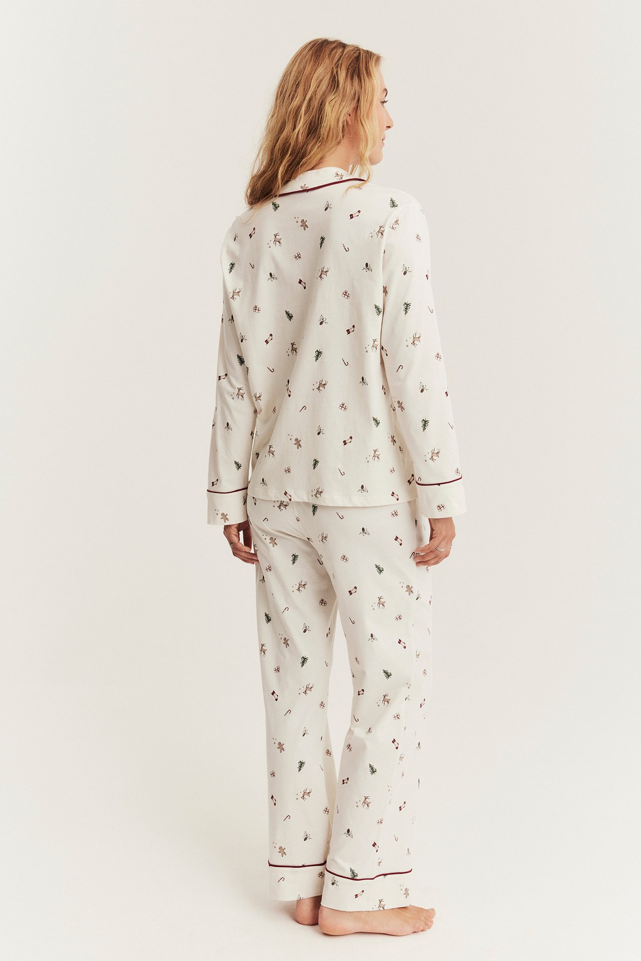 Naisen takakuva kermanvärisessä jouluaiheisessa pyjamasetissä, jossa on viininpunainen reunanauha.