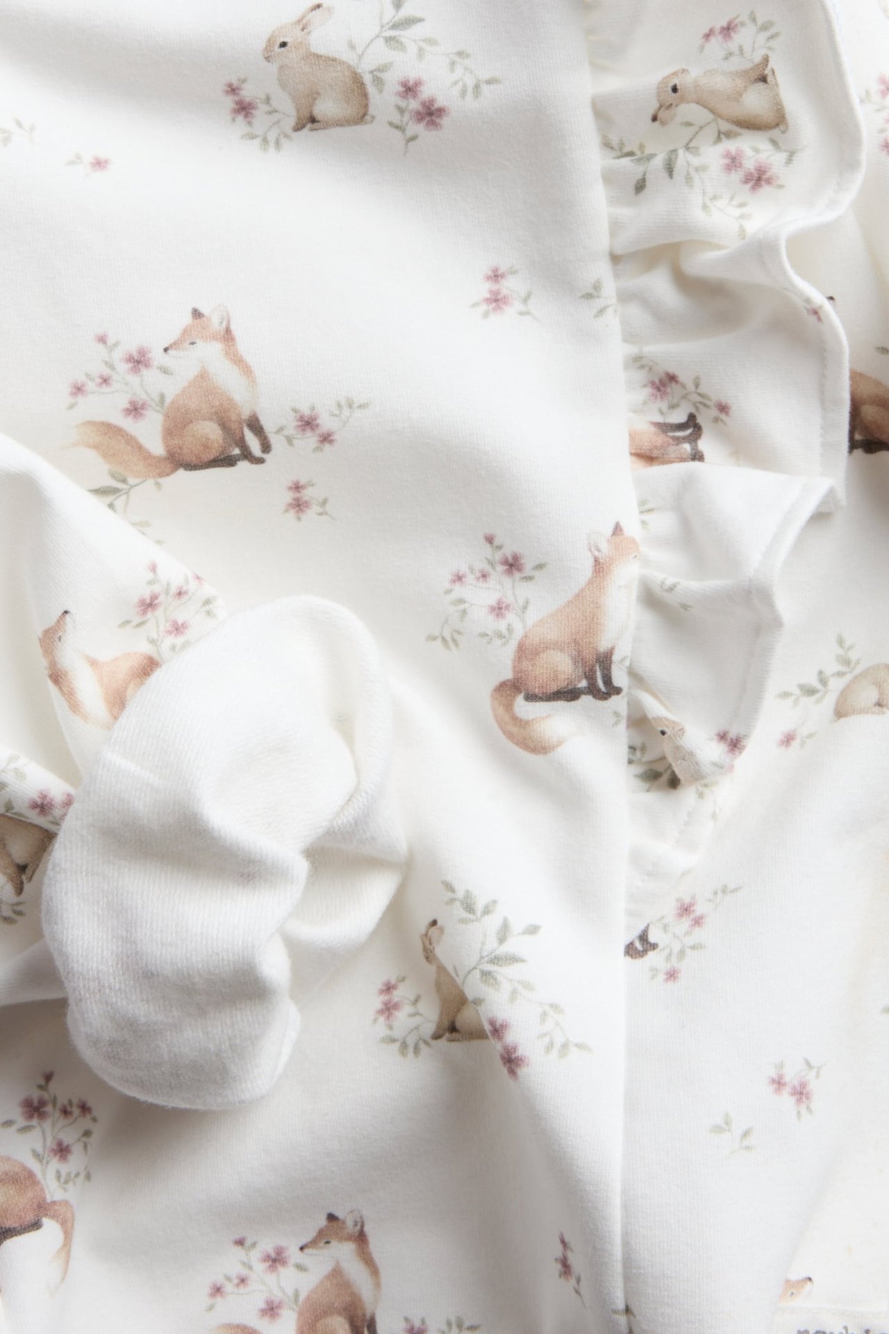 Närbild av vitt babyplagg med räv- och kanintryck och volangdetalj, bredvid en vit babysocka.