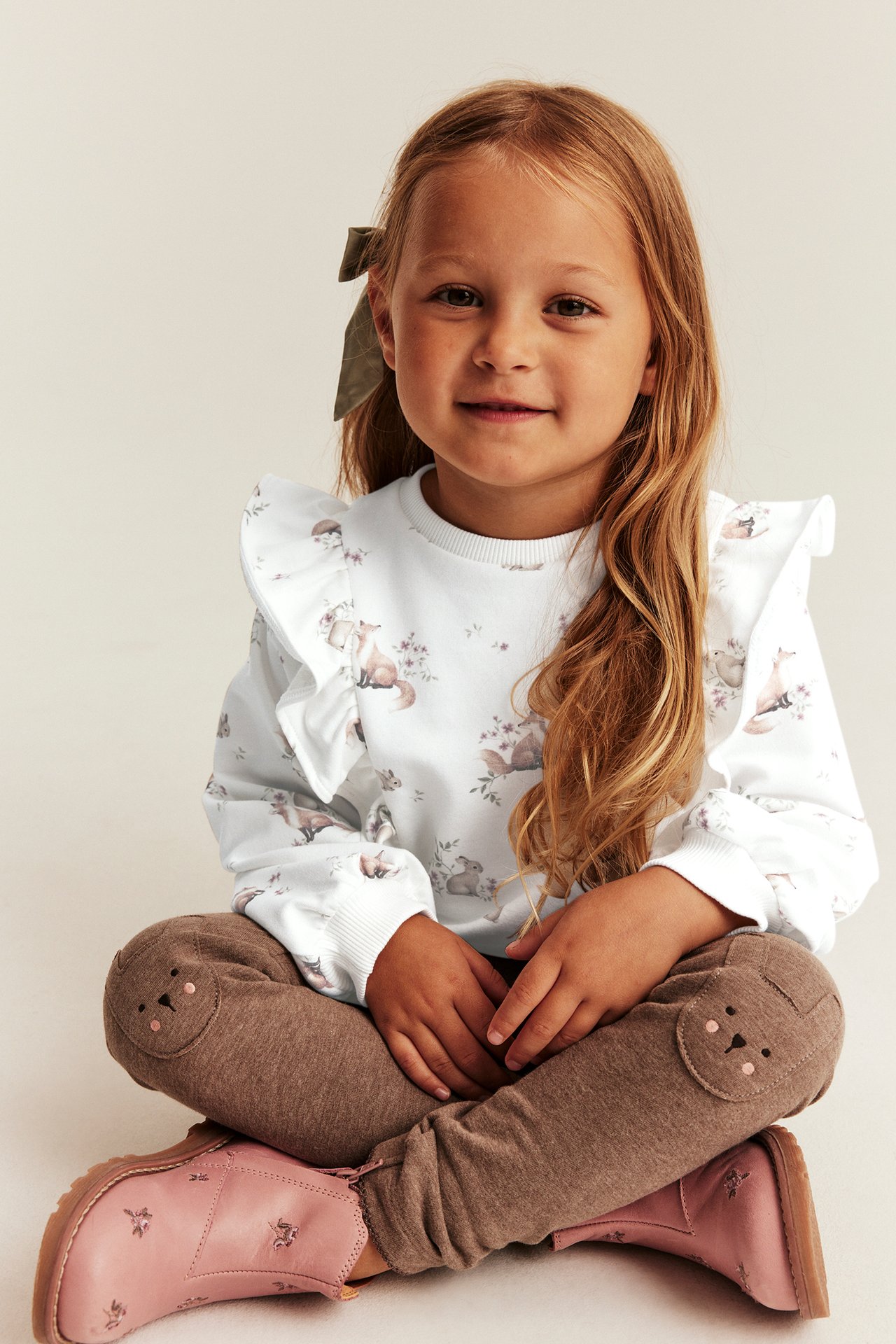 Barn i hvit volanggenser med dyreprint, brun leggings med dyreknelapper, rosa ankelstøvler.