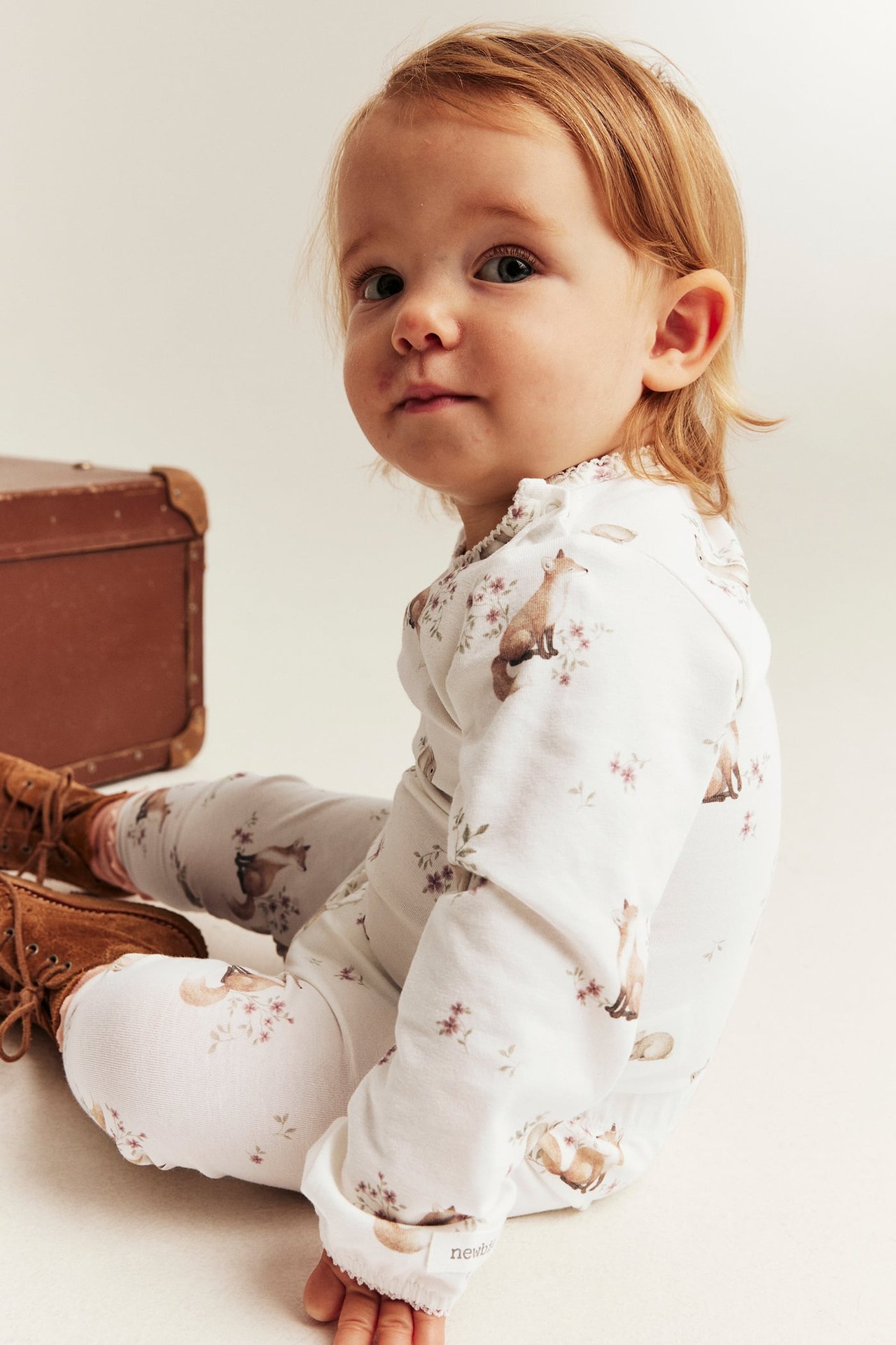 Frontbilde av baby i hvit langermet jumpsuit med rev- og blomstermønster og brune snørestøvler.