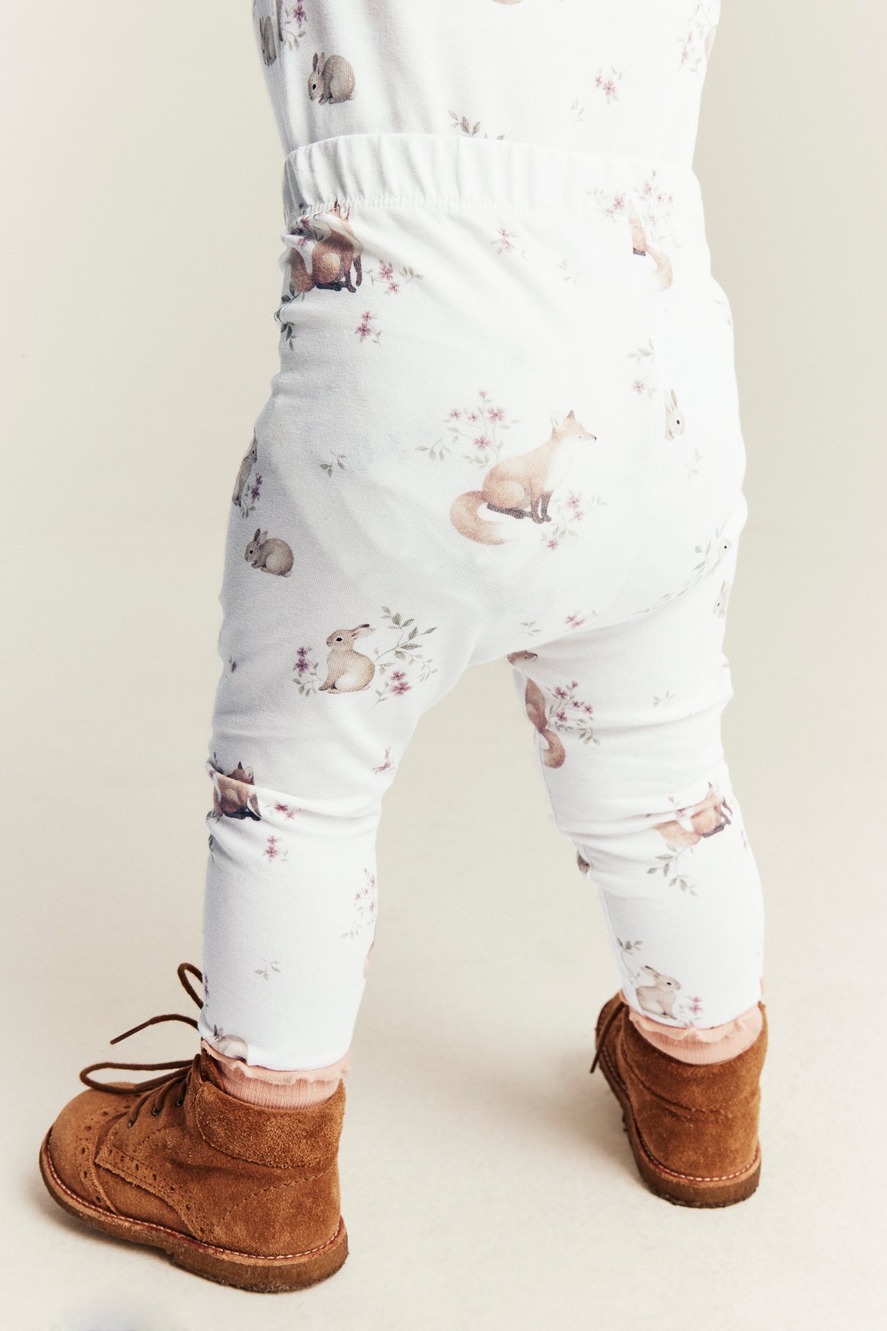 Bakfra: baby i hvite leggings med dyremønster og brune snørestøvler.