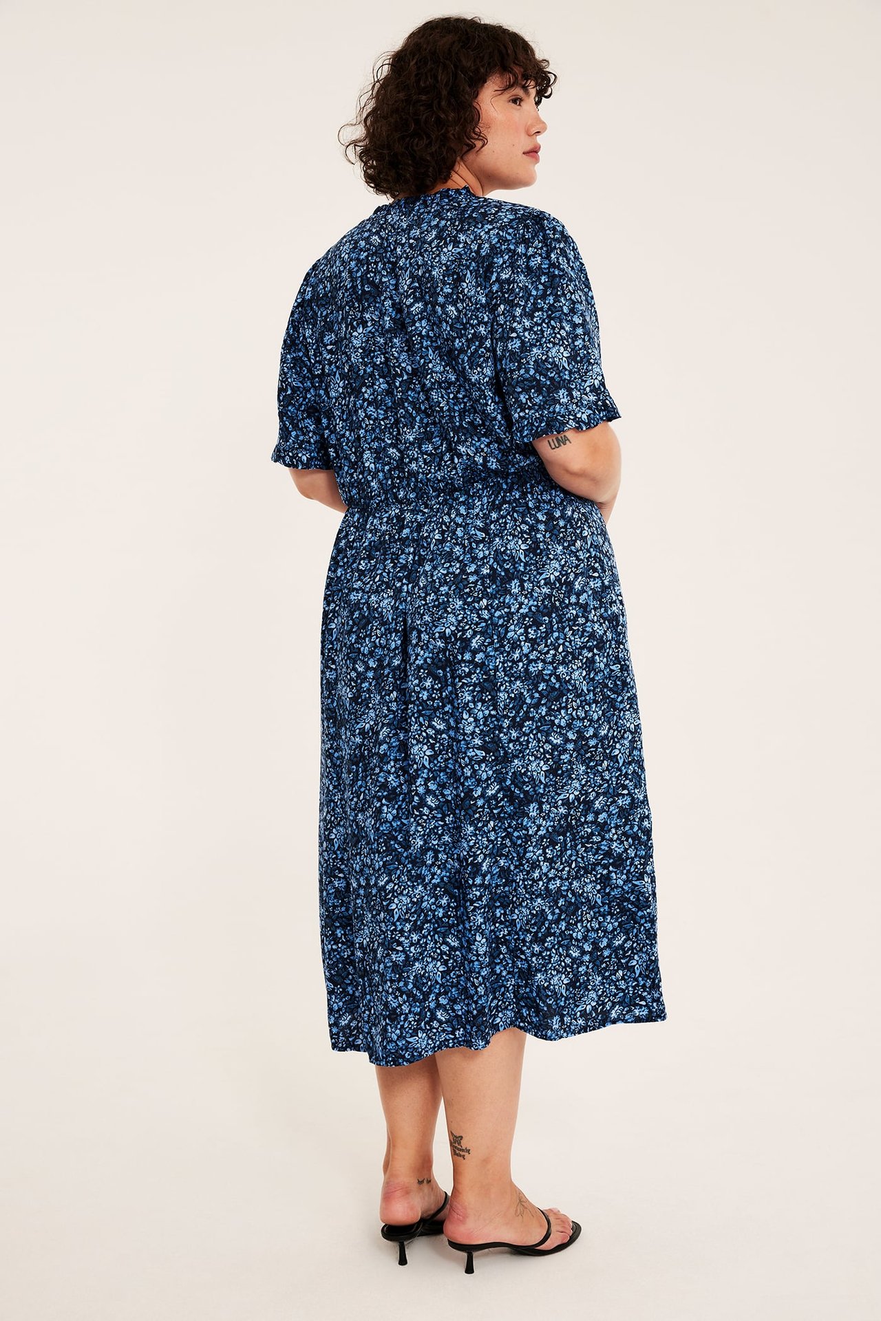 Baksidebilde av en kvinne i en blå blomstret midikjole og svarte sandaler med hæl.