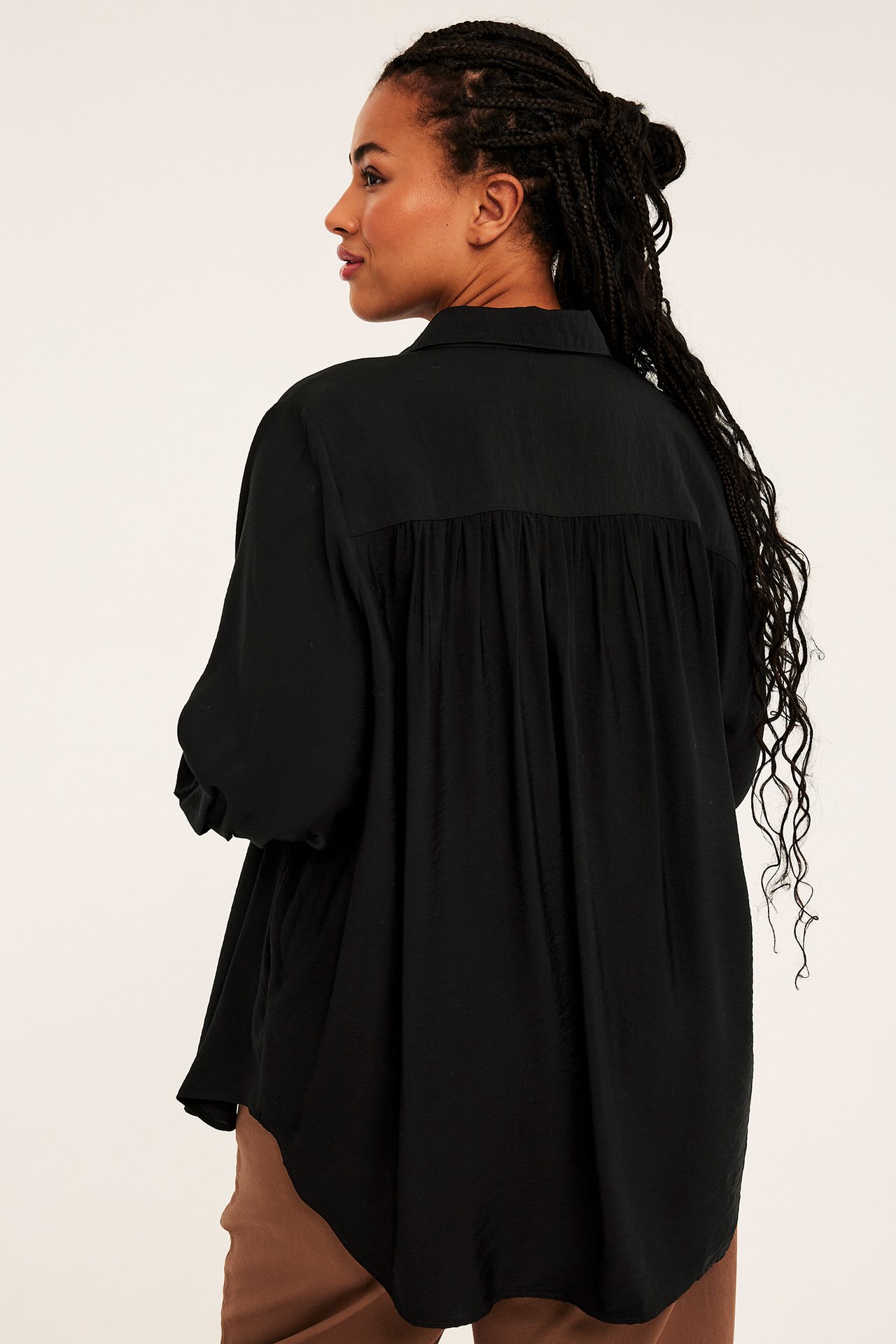 Baksidevisning av kvinnelig modell iført en oversized svart bluse med rynket rygg og brune bukser.