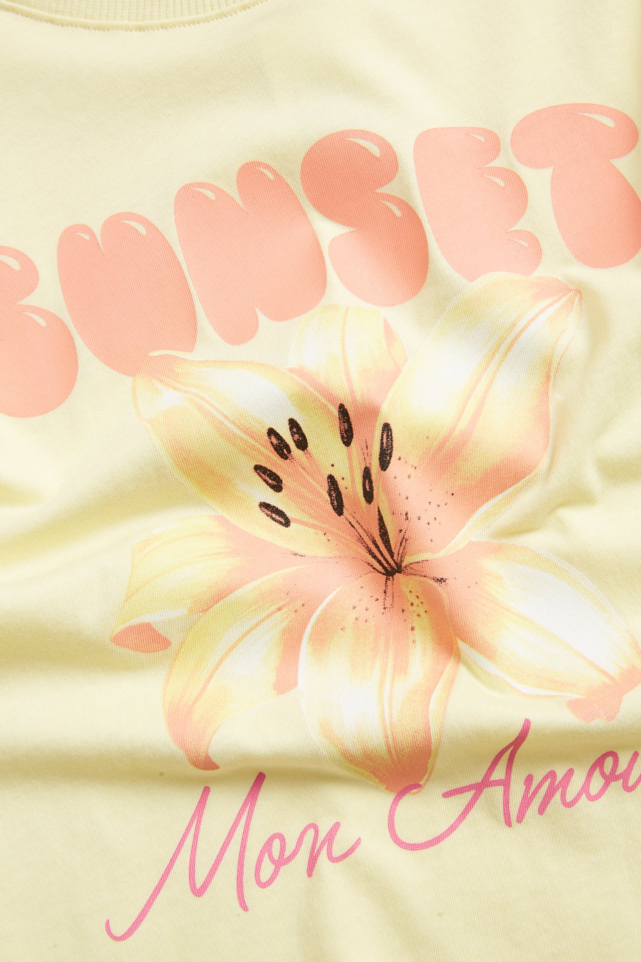 Zbliżenie: jasnożółta bawełniana koszulka z różowym kwiatowym nadrukiem i tekstem.