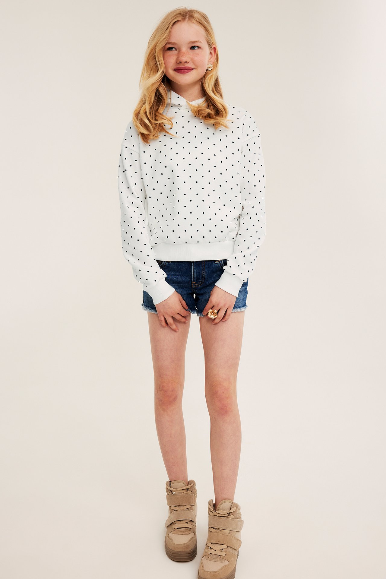 Barn forfra, iført hvit prikkete hettegenser, mørkeblå denimshorts og beige høye joggesko.