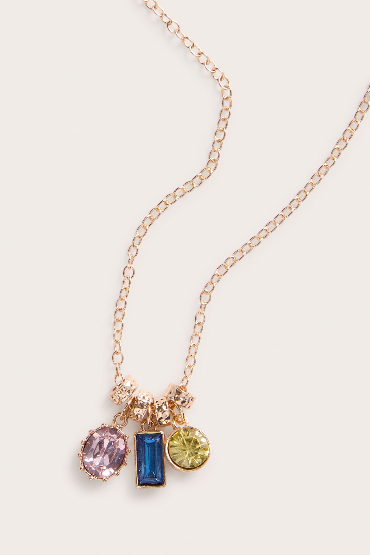 Närbild av ett guldfärgat halsband med tre färgglada ädelstensberlocker: rosa oval, blå rektangel, gul rund.