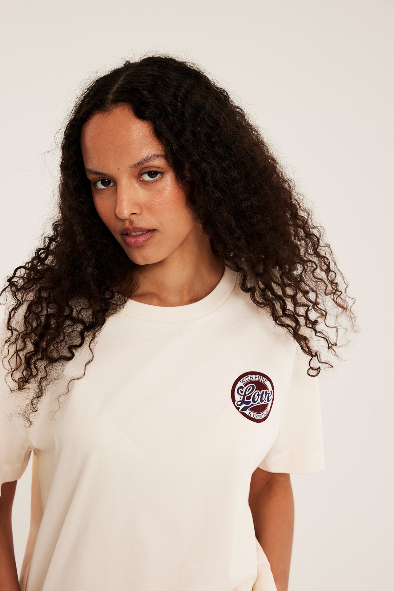 Frontvy av en kvinna i en offwhite bomulls-T-shirt med ett cirkulärt 'Love'-tryck på bröstet.
