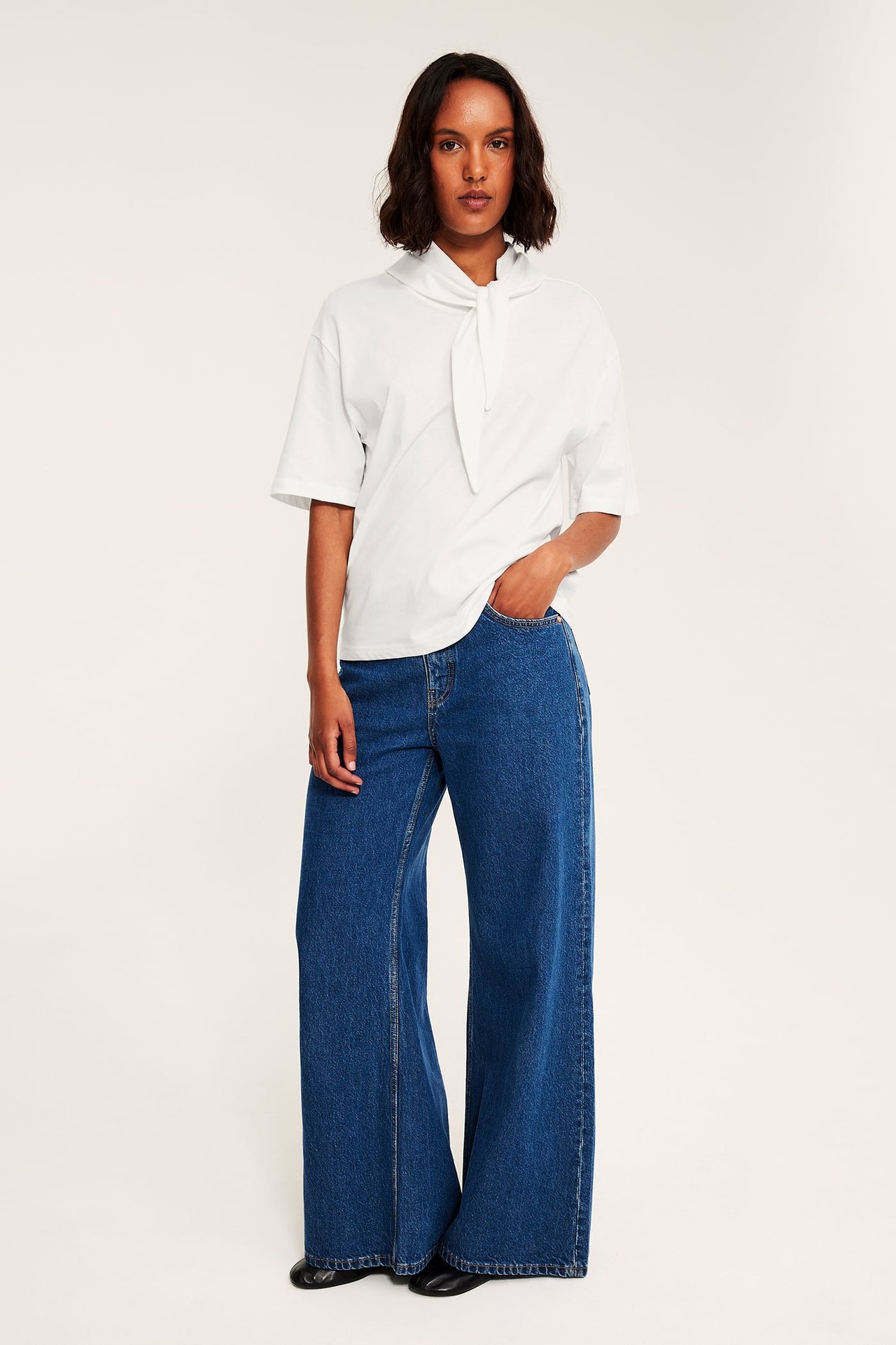 Framifrån kvinnlig modell i vit kortärmad topp med knytdetalj, blå vida jeans, svarta platta skor.