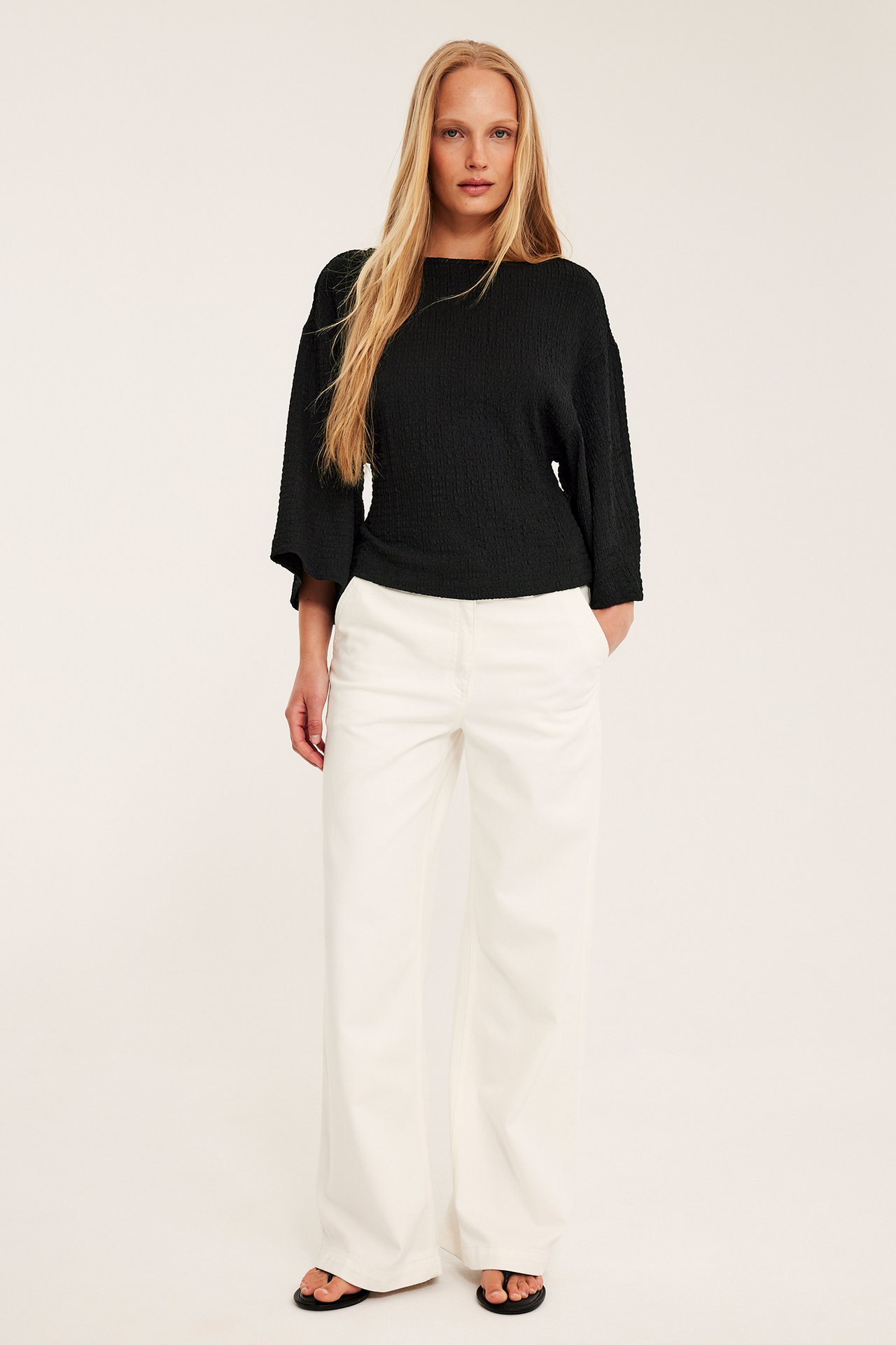 Nainen edestä: musta kuviollinen toppi leveillä kimono-hihoilla, valkoiset leveälahkeiset housut, mustat varvassandaalit.