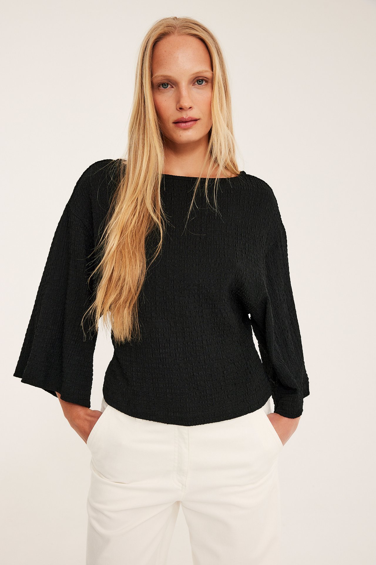 Nainen edestä mustassa, strukturoidussa topissa, jossa leveät kimonohihat, ja valkoisissa housuissa.