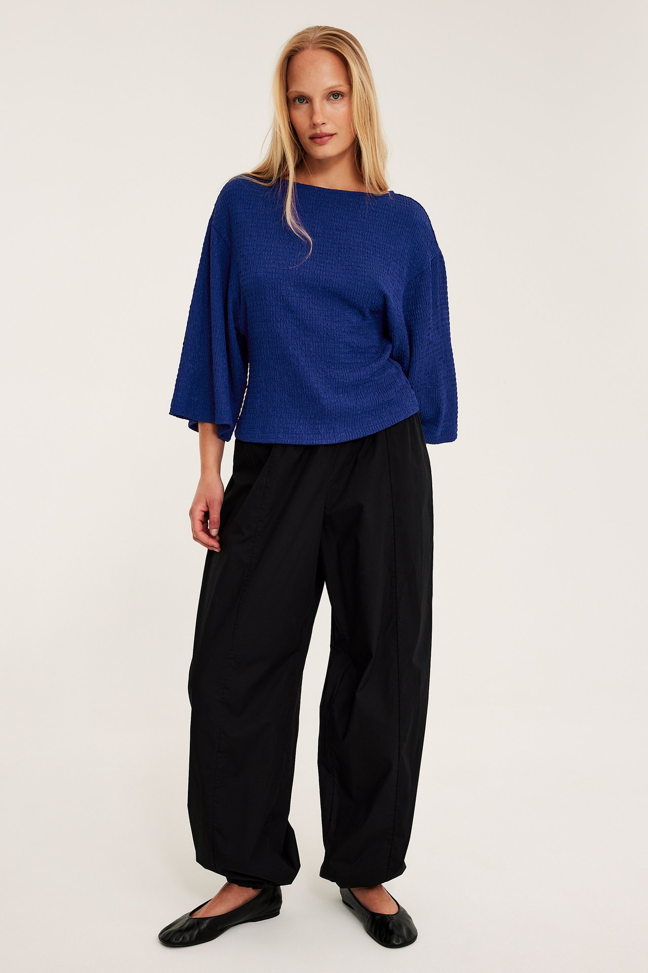 Nainen sinisessä strukturoidussa topissa, leveät kimono-hihat. Mustat housut ja kengät.