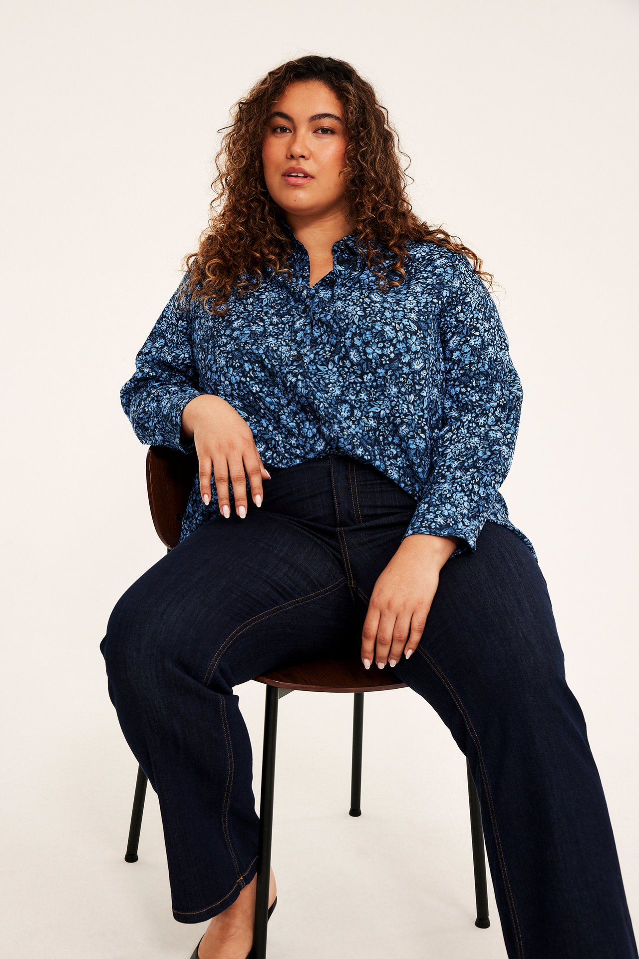 Framifrån: kvinna i blå blommig långärmad skjorta, mörkblå jeans och svarta klackskor.