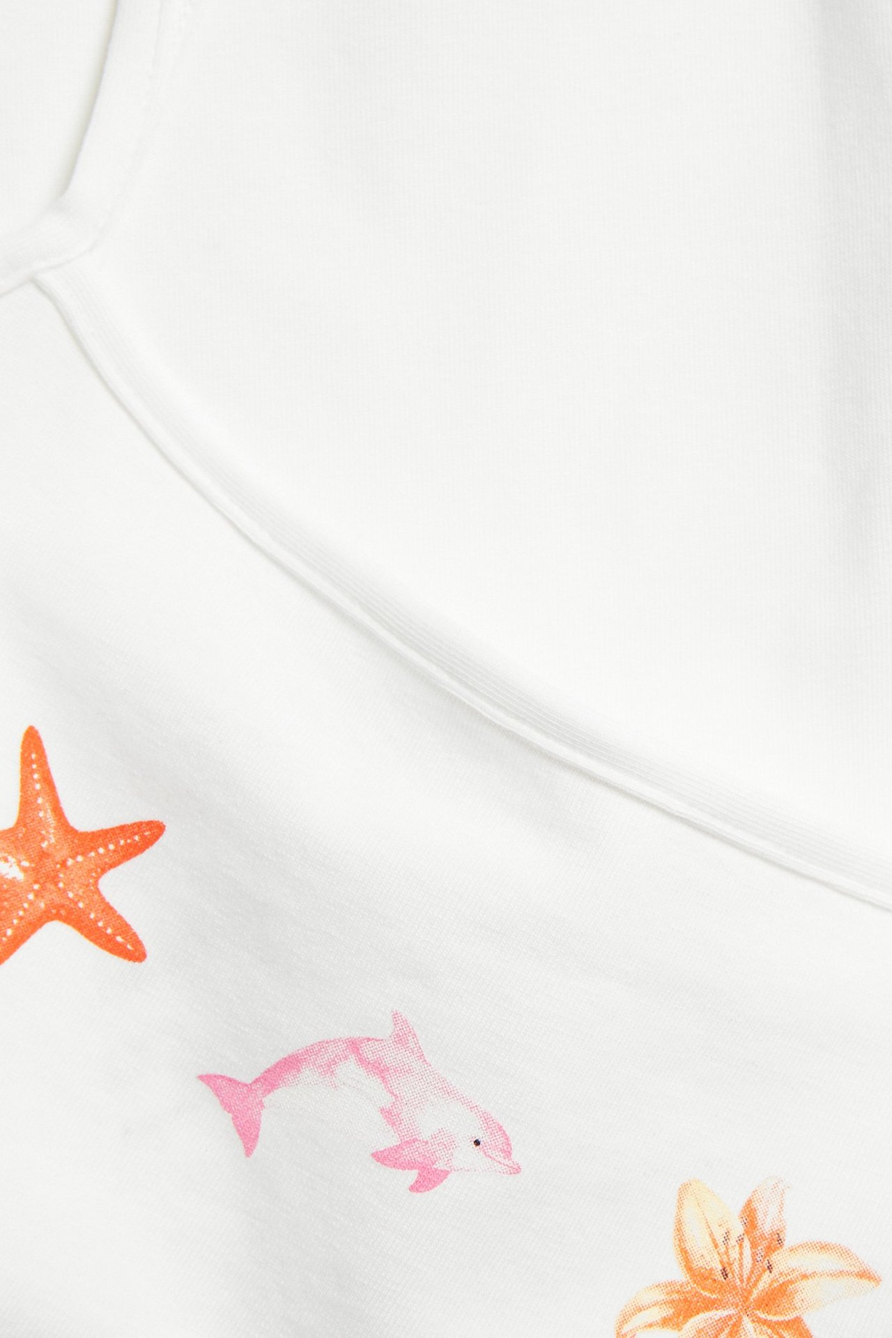 Zbliżenie na kremowy top z domieszką bawełny z nadrukiem różowego delfina, pomarańczowej rozgwiazdy i kwiatu.