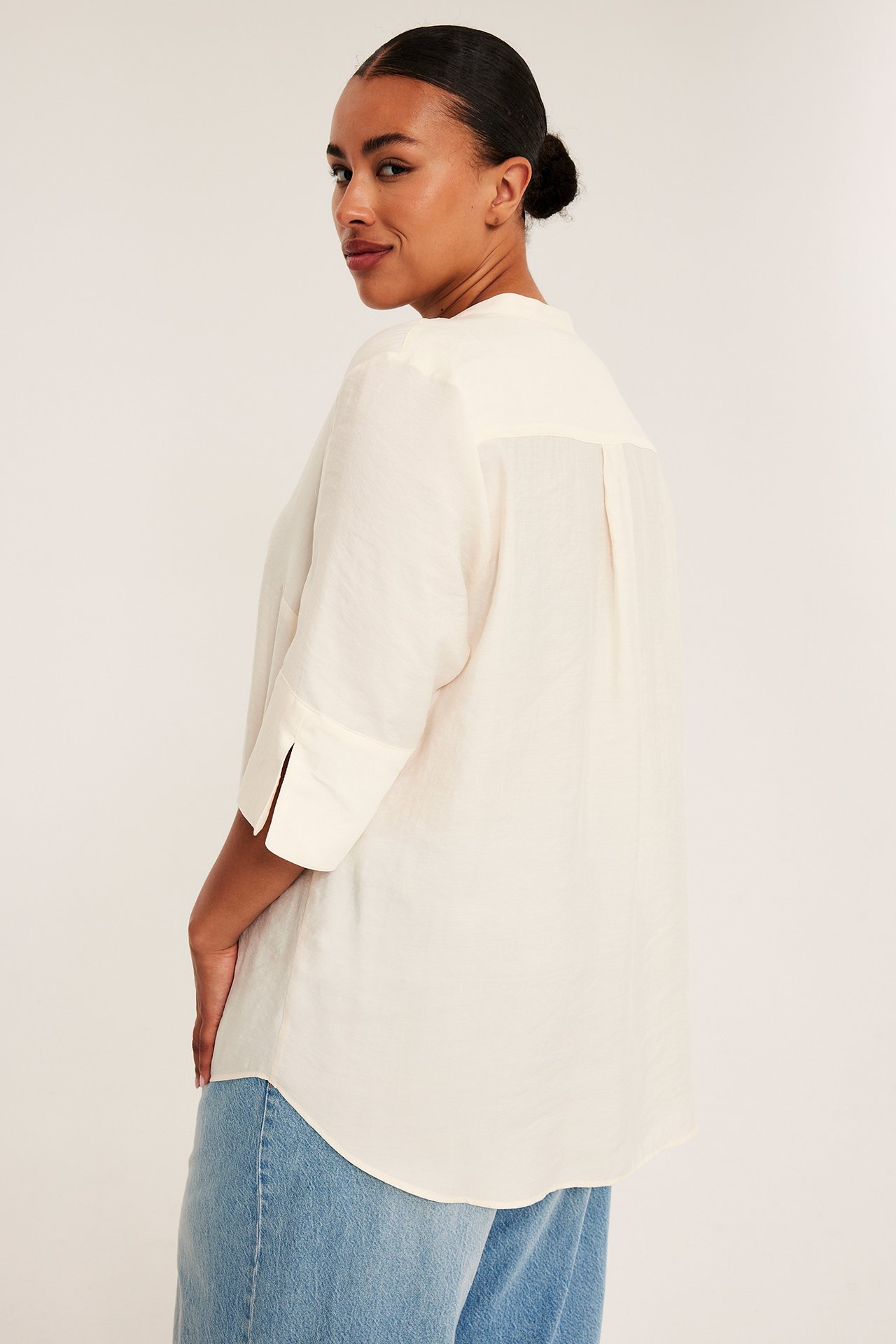 Sidebilde av kvinne i en beige oversized skjorte med 3/4 ermer og lyseblå jeans.