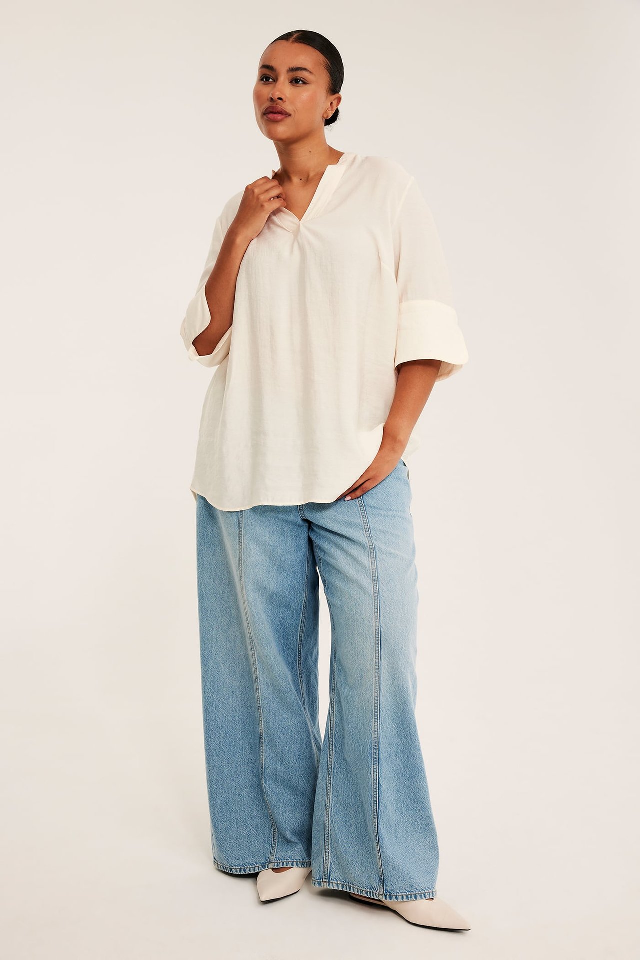 Framifrån: kvinna i offwhite V-ringad tunika, ljusblå jeans med vida ben, och beige platta skor.