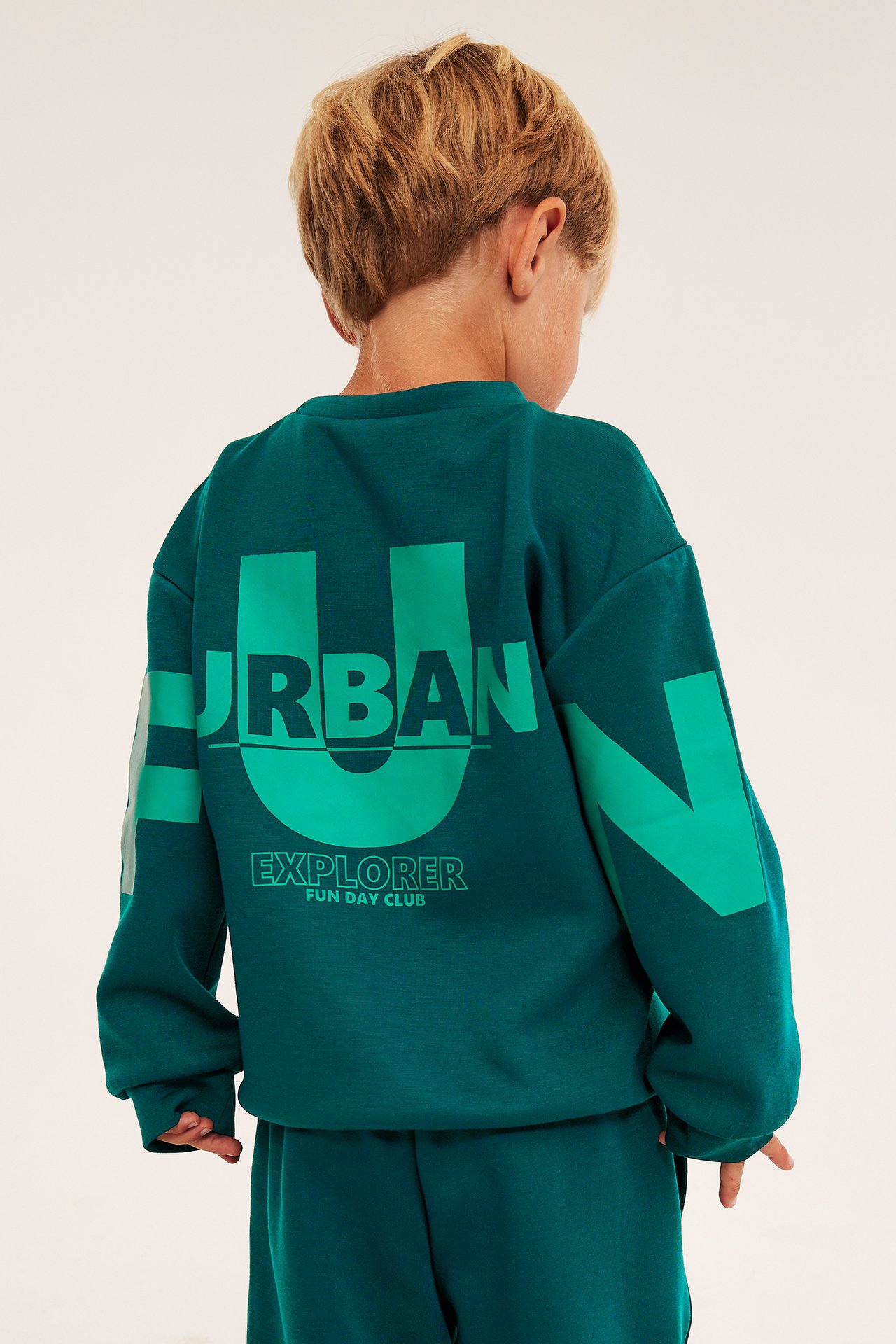 Lapsi takaapäin tummanvihreässä collegepaidassa, jossa vaaleanvihreä 'URBAN EXPLORER' -printti.