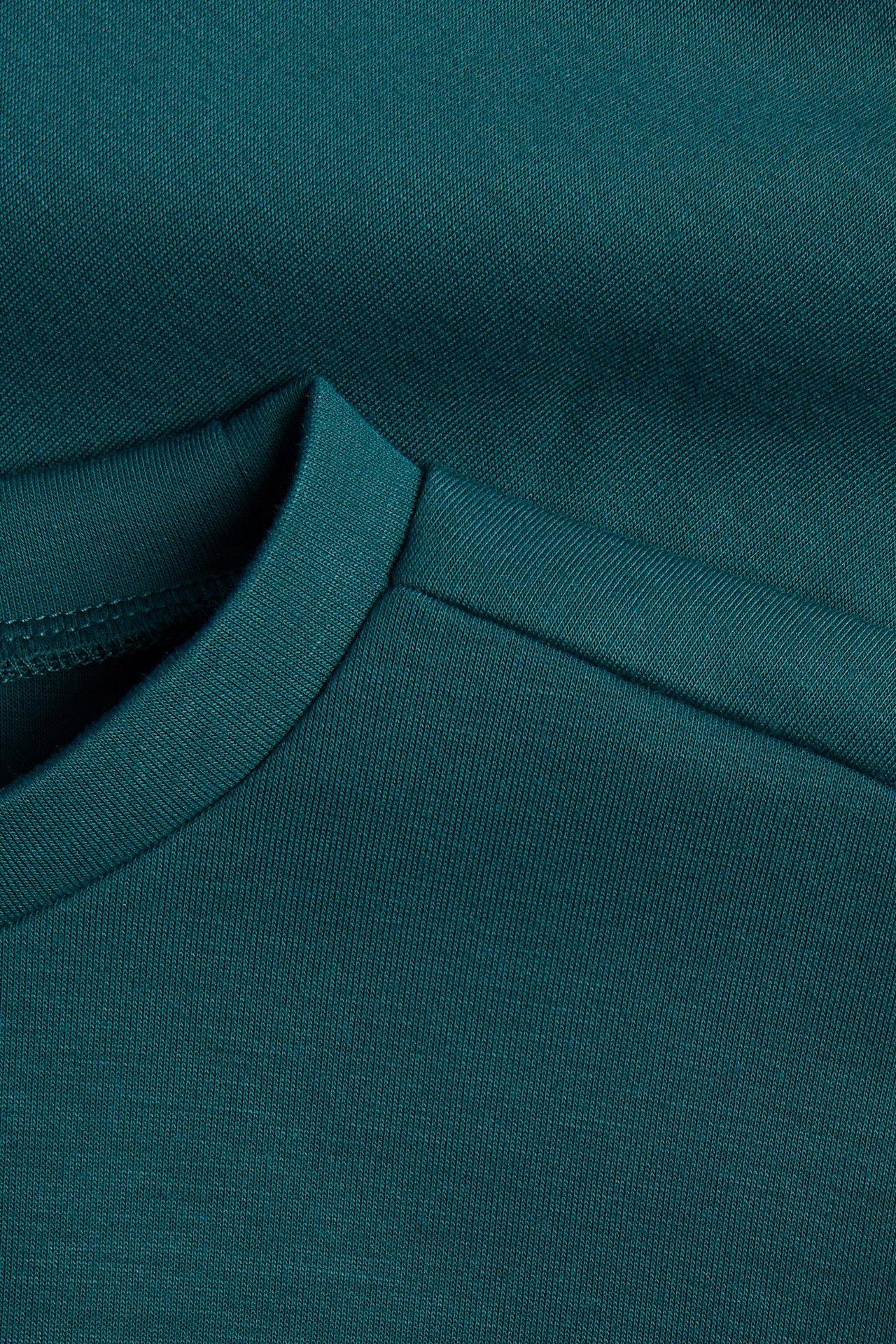Närbild på en mörk turkos t-shirt i bomullsjersey med ribbad rund halsringning och axelsöm.