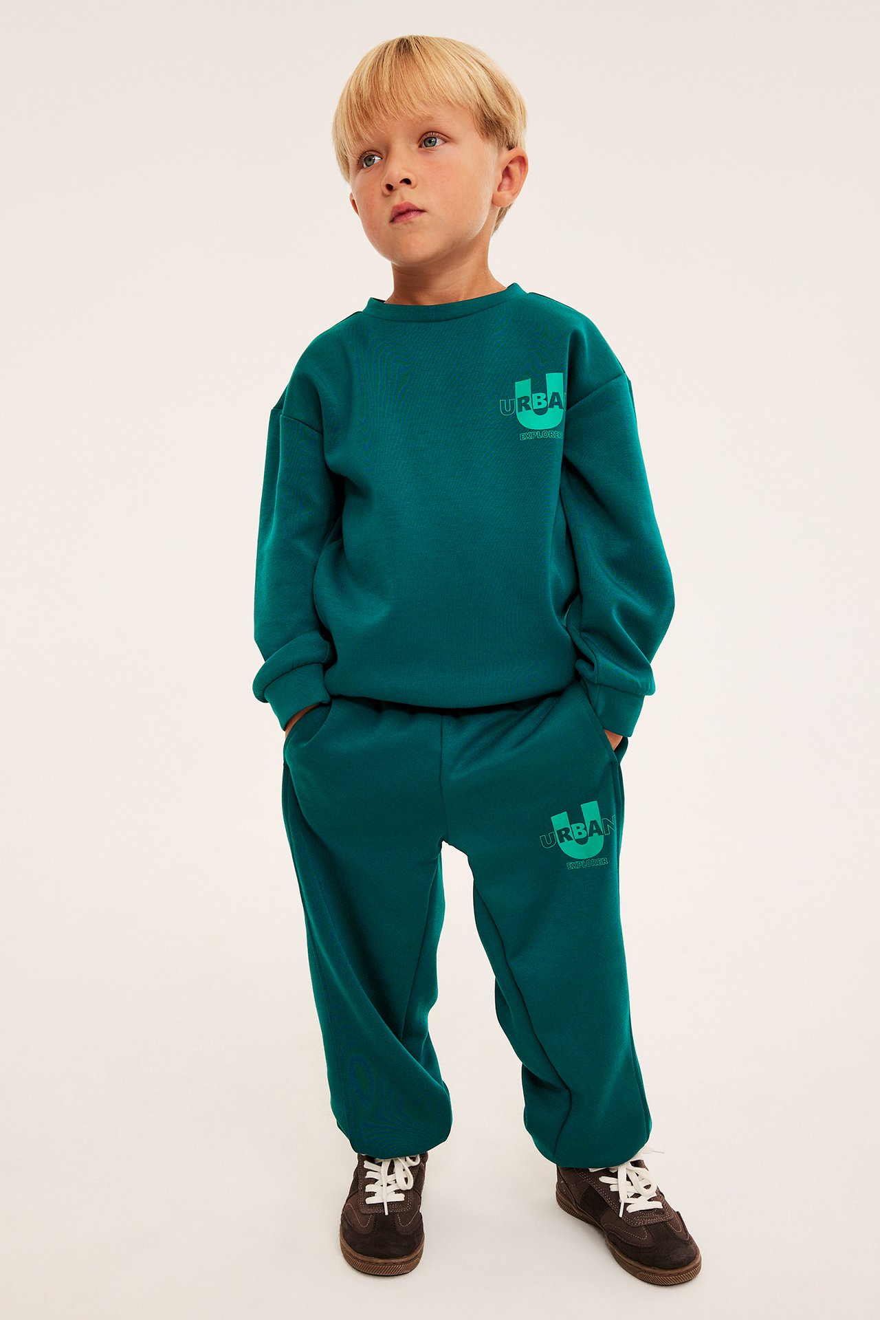 En pojke i mörk turkos URBA-mönstrad sweatshirt och matchande mjukisbyxor med bruna sneakers, framifrån.