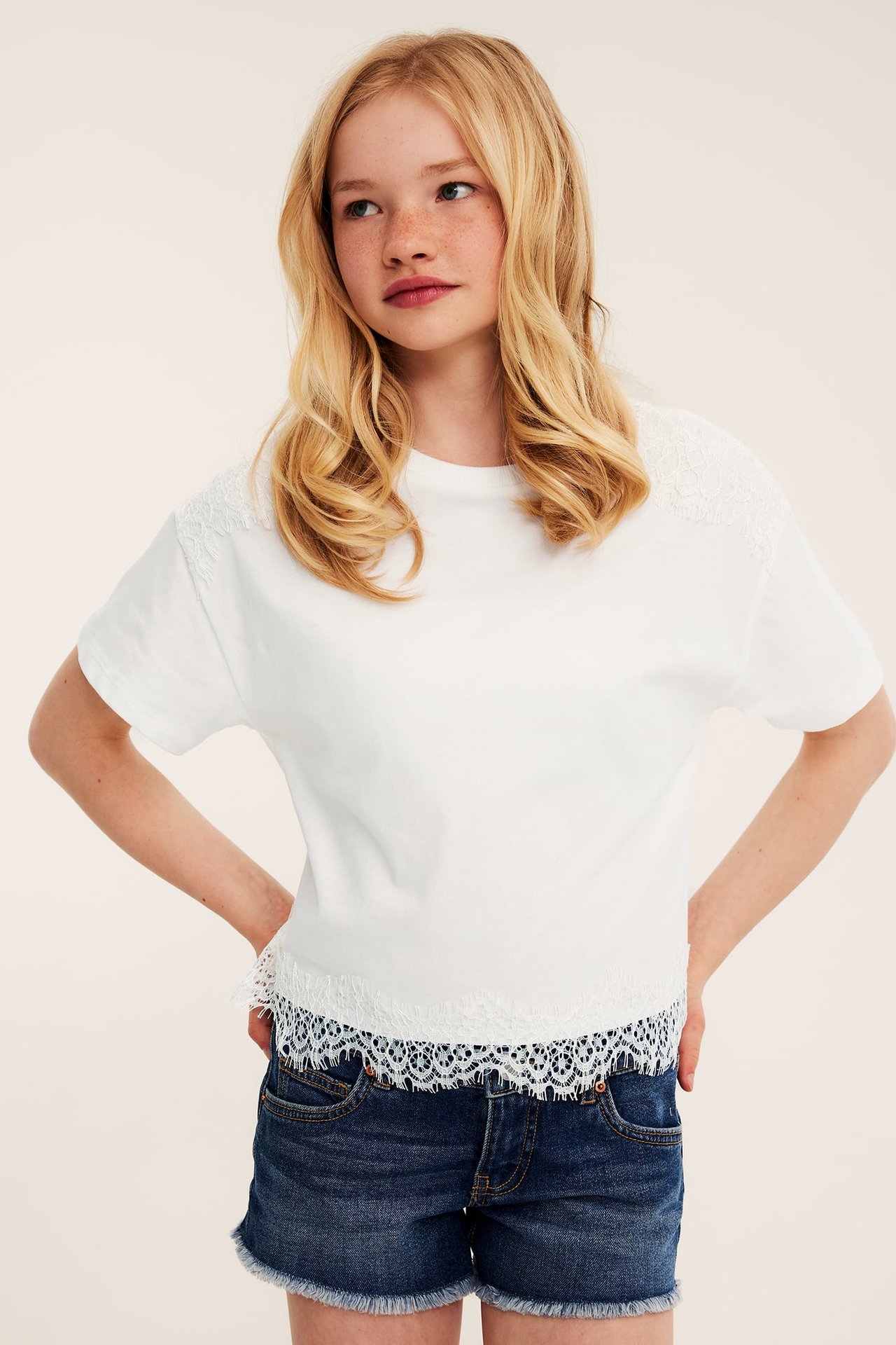 Framsida: en flicka i offwhite T-shirt med spetsdetaljer på axlar och nederkant, samt mörkblå denimshorts.