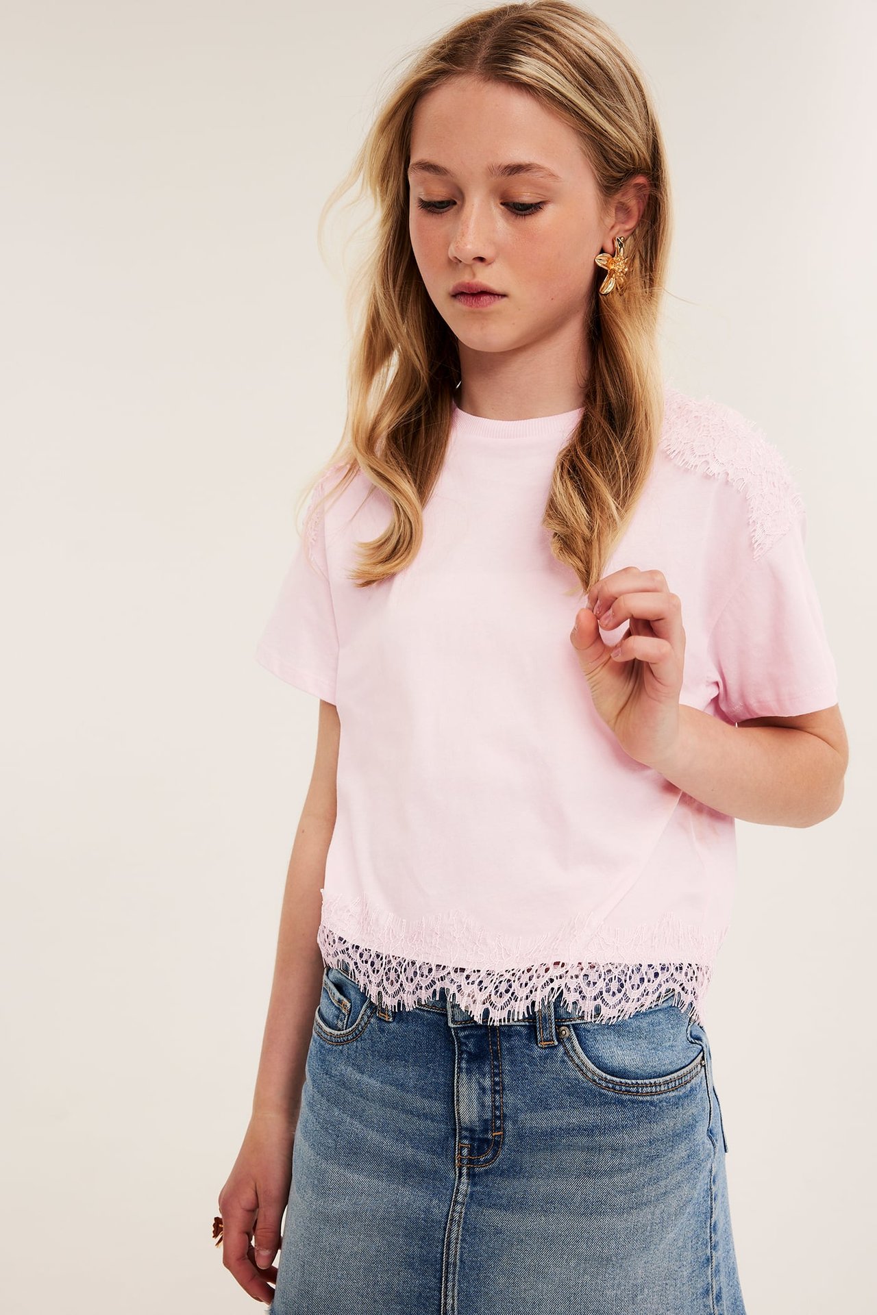 Framifrån, ett barn i ljusrosa T-shirt med spetsdetaljer, blå jeanskjol och guldfärgade sjöstjärneörhängen.
