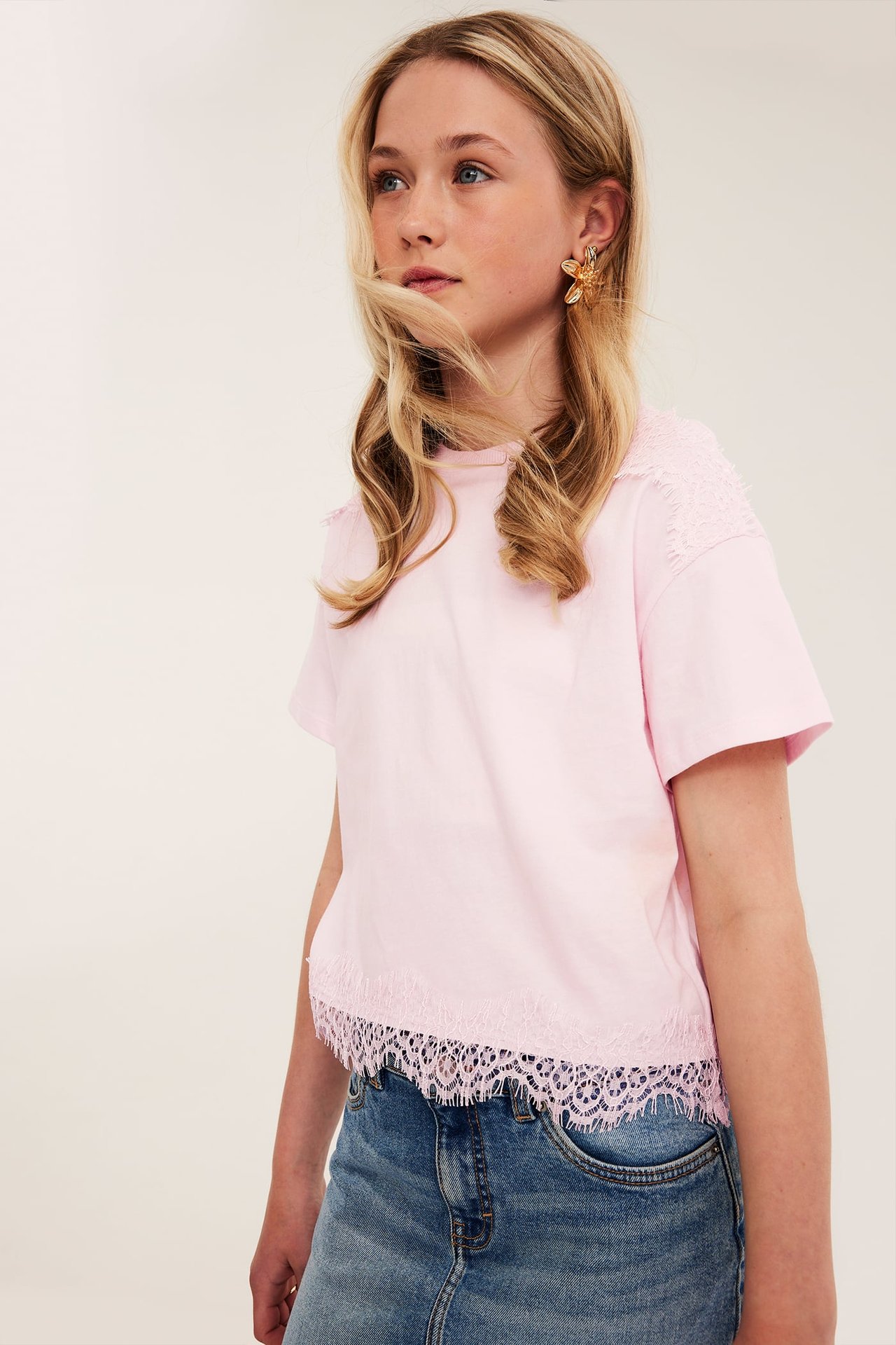 Barn bär en ljusrosa T-shirt med spetsdetaljer, en blå jeanskjol och ett guldfärgat blommigt örhänge.